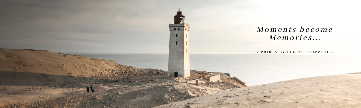 A tall lighthouse on a sandy beach with two people walking nearby, overcast sky in the background, with overlay text 'Moments become Memories... - Prints by Claire Droppert'.