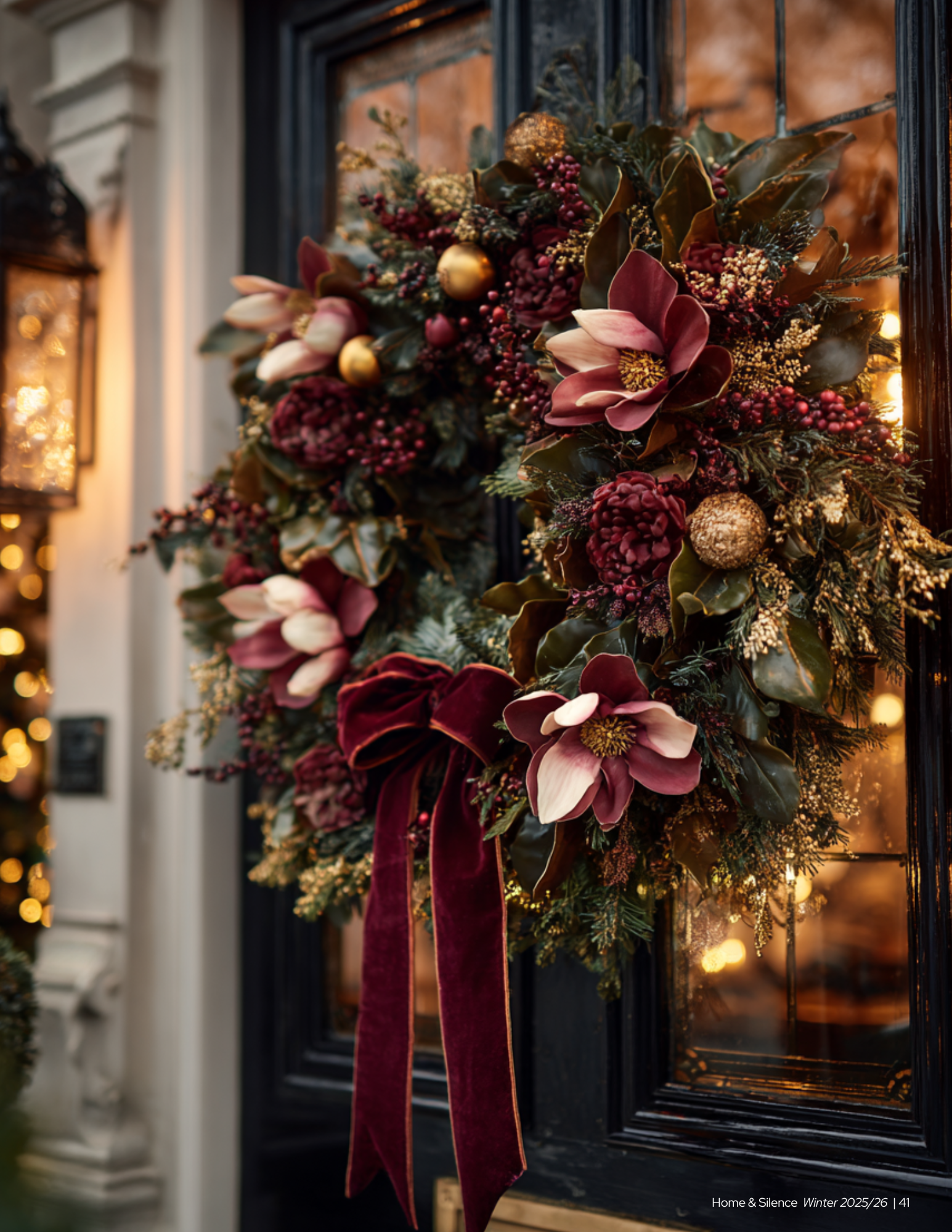 A festive Christmas wreath with pink, burgundy, and gold ornaments, holly leaves, magnolia flowers, pinecones, and a maroon velvet ribbon, hanging on a black door with warm lighting in the background.