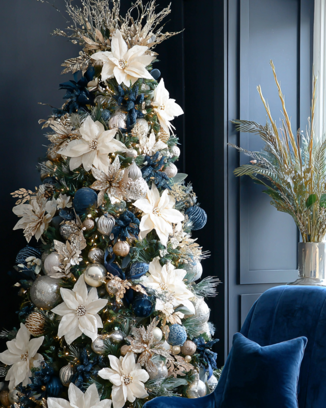 Elegant Christmas tree decorated with navy blue and gold ornaments, ribbons, and florals in a dark, moody living room.
