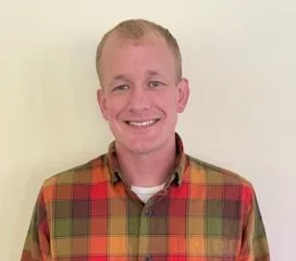A smiling man with short blonde hair wearing a red and yellow plaid shirt against a plain background.