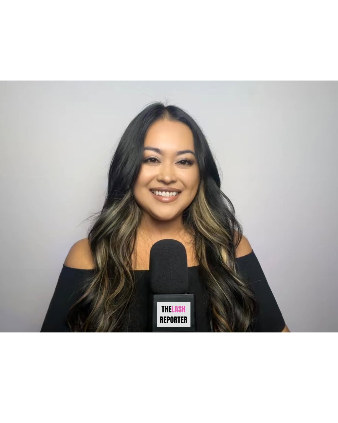 A woman with long wavy hair, wearing an off-the-shoulder black top, smiling, standing in front of a plain light gray background, holding a microphone with a label that reads 'The Lash Reporter'.