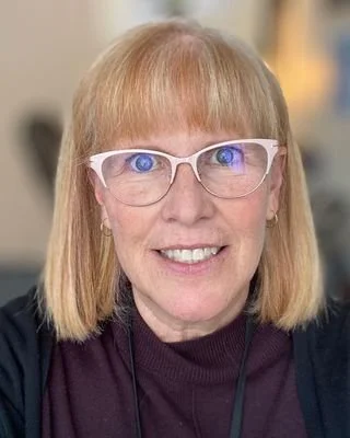 A woman with blonde hair and blue eyes wearing white glasses and a dark top, smiling for a photo.
