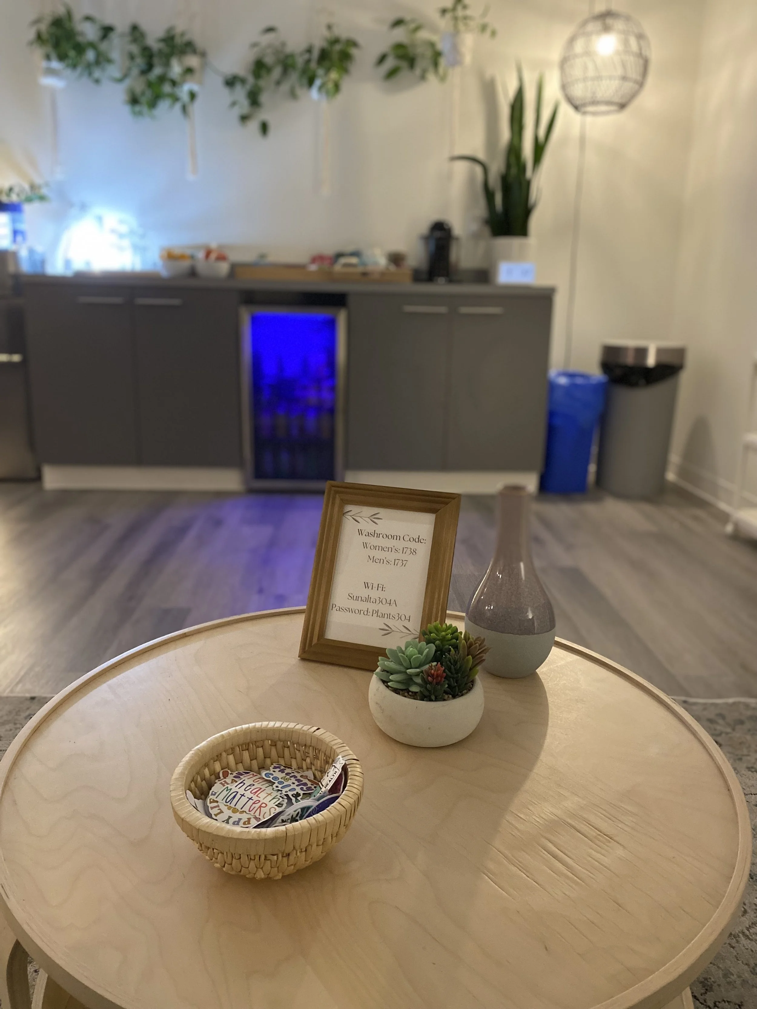 Close-up of a round wooden table with a small basket containing colorful cards and pens, a framed sign displaying washroom codes and Wi-Fi details, a gray vase, and a small potted succulent plant in an indoor setting with gray cabinets, a blue-lit mini fridge, and hanging woven lamps in the background.