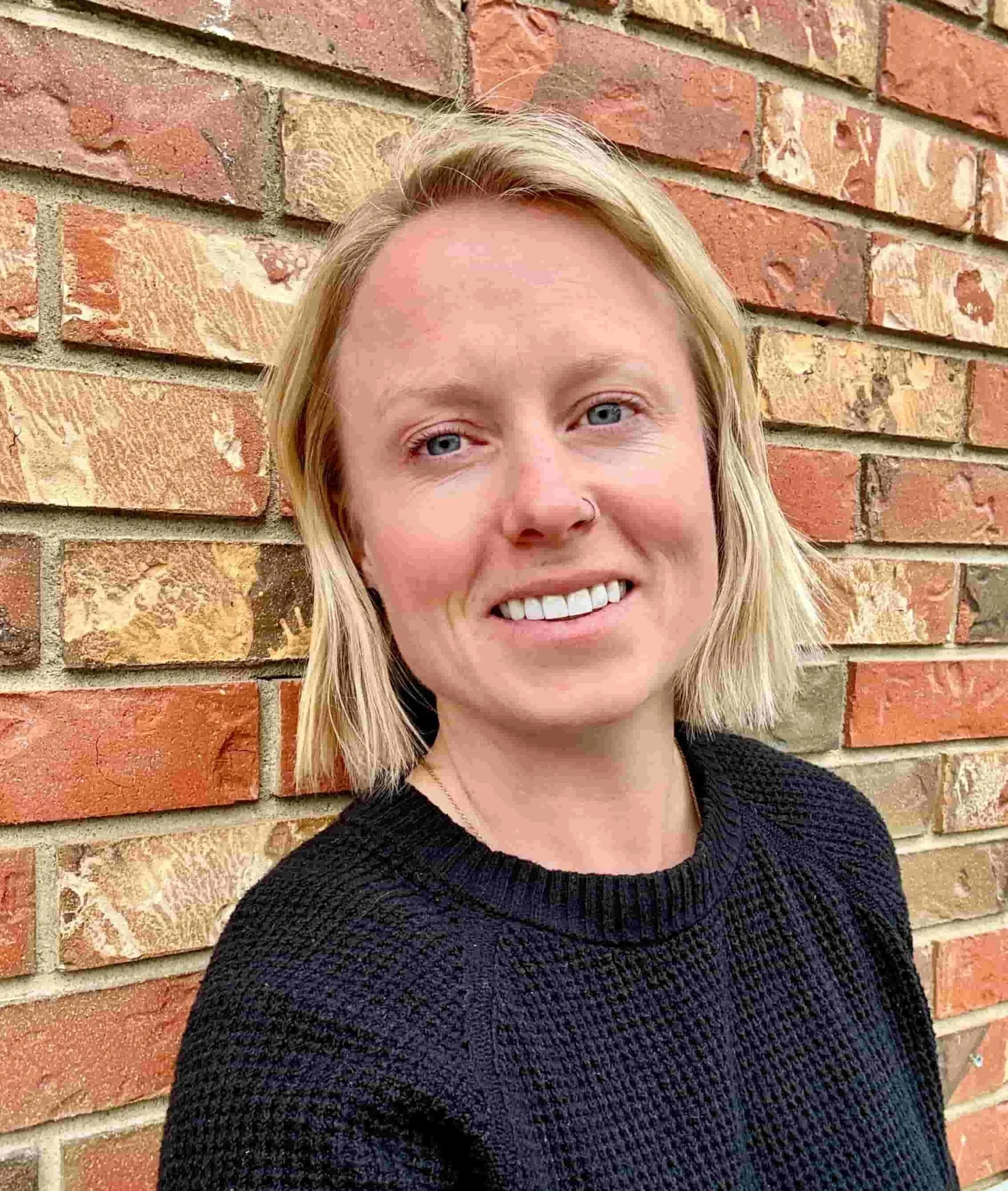 A woman with short blonde hair, light skin, wearing a black textured sweater, smiling, standing outdoors in front of a multicolored brick wall.
