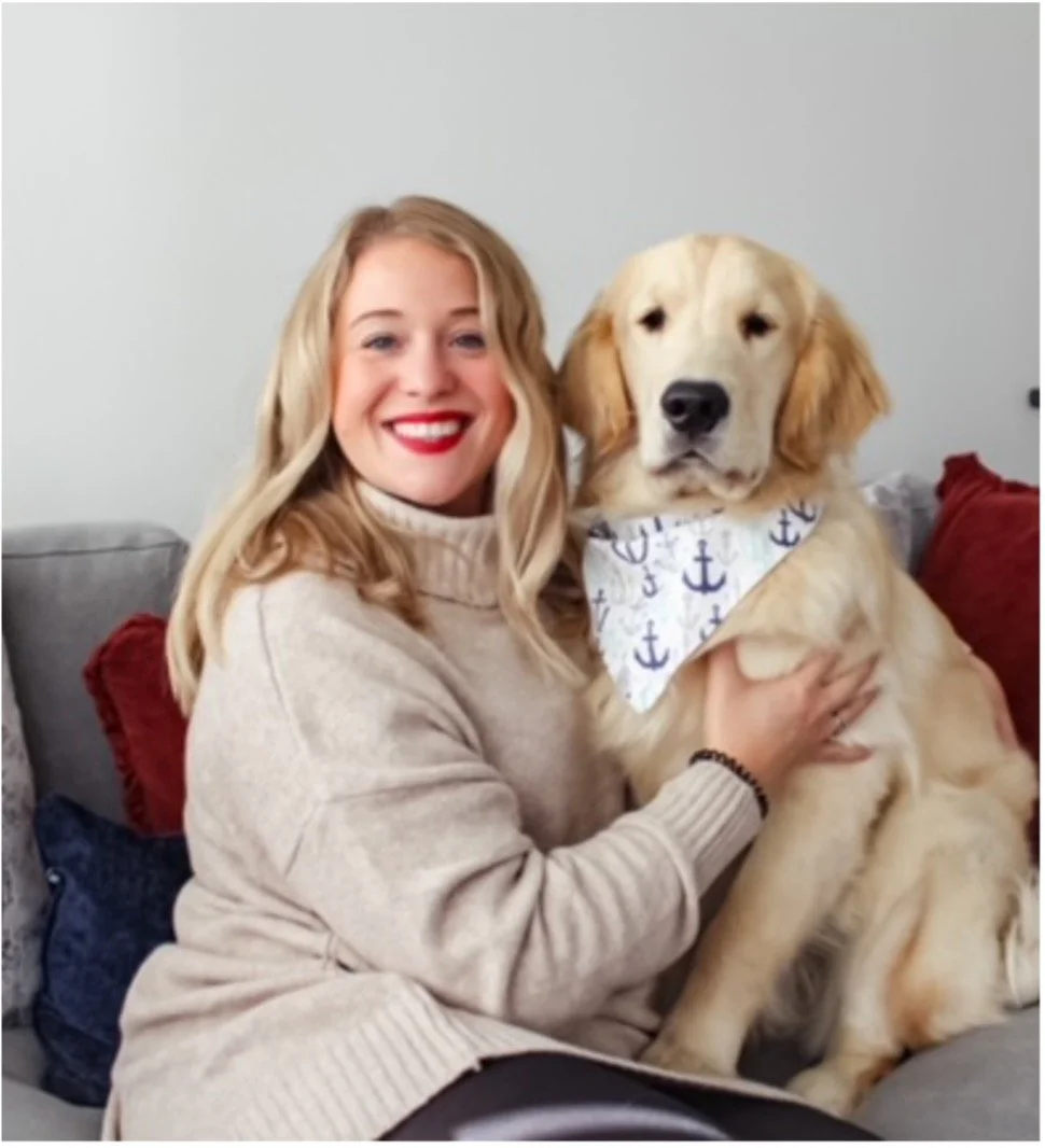 A woman with blonde hair, wearing a beige sweater, sitting on a couch with red and gray cushions, holding a large golden retriever with a white bandana decorated with blue anchors.