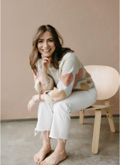 A woman sitting on a wooden chair, smiling, with long brown hair, wearing a multicolored sweater and white pants in a minimalistic room.