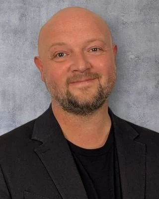 Headshot of a man with a bald head and beard, wearing a black blazer and black shirt, against a gray background.