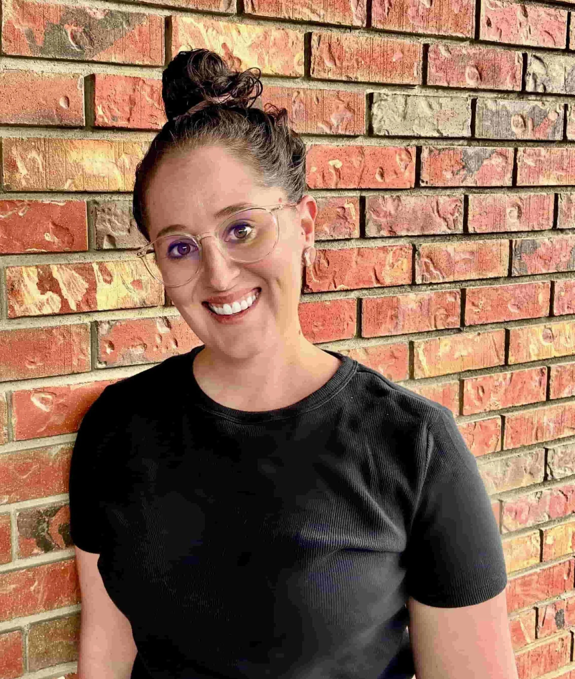 A young woman with glasses and a top bun hairstyle, smiling and wearing a black t-shirt, standing against a brick wall.