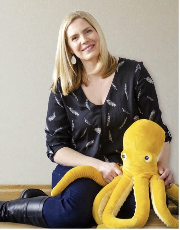 A woman with blonde hair, wearing a black blouse with feather prints, sitting on the floor and smiling while holding a large yellow plush octopus.
