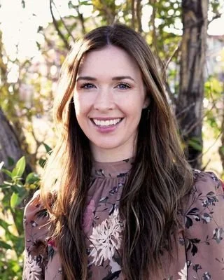 A young woman with long brown hair and a bright smile standing outdoors with trees and sunlight in the background.