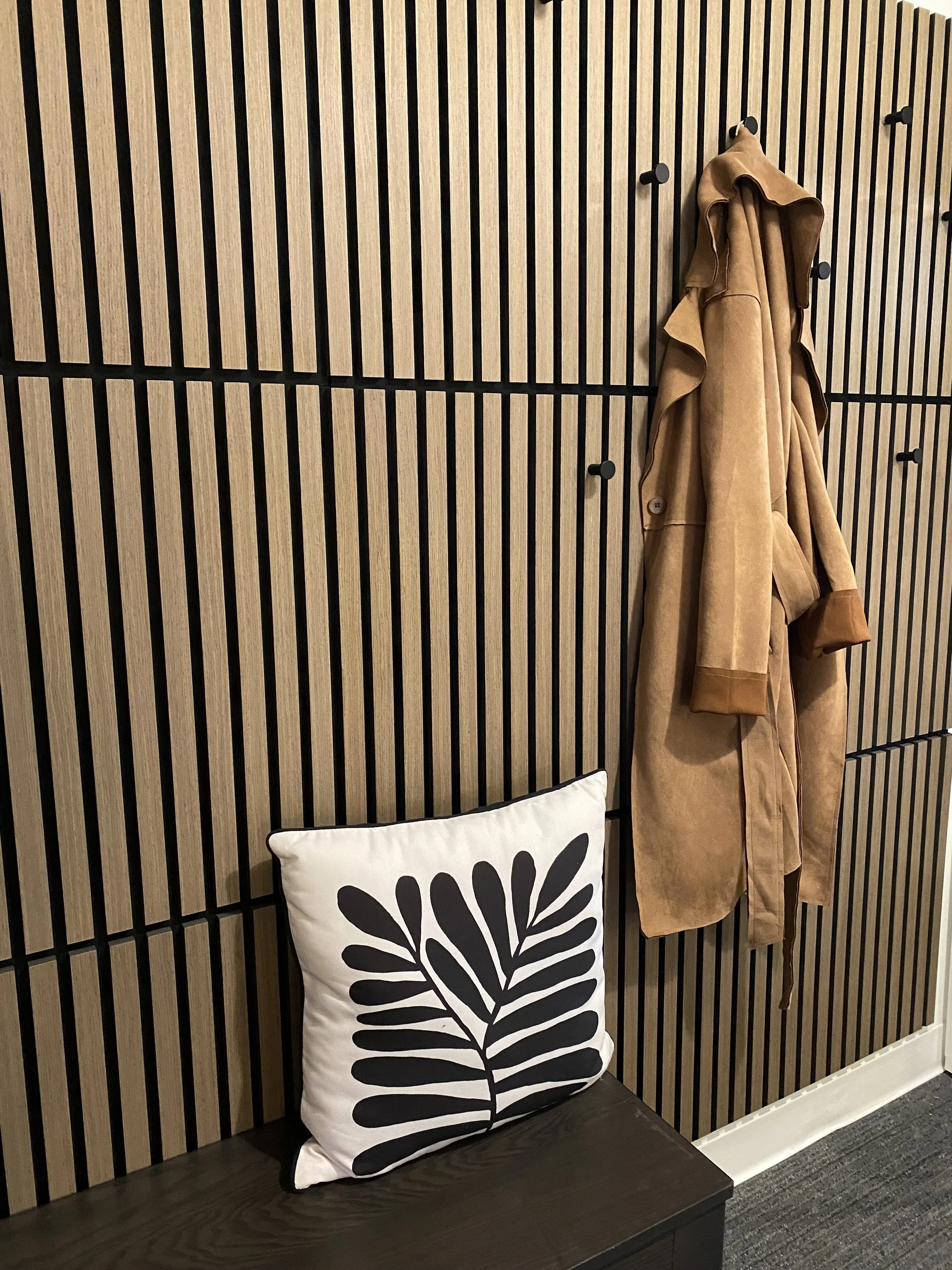 A coat hanging on a wall with vertical wooden slats, above a bench with a black and white decorative pillow depicting a stylized leaf design.