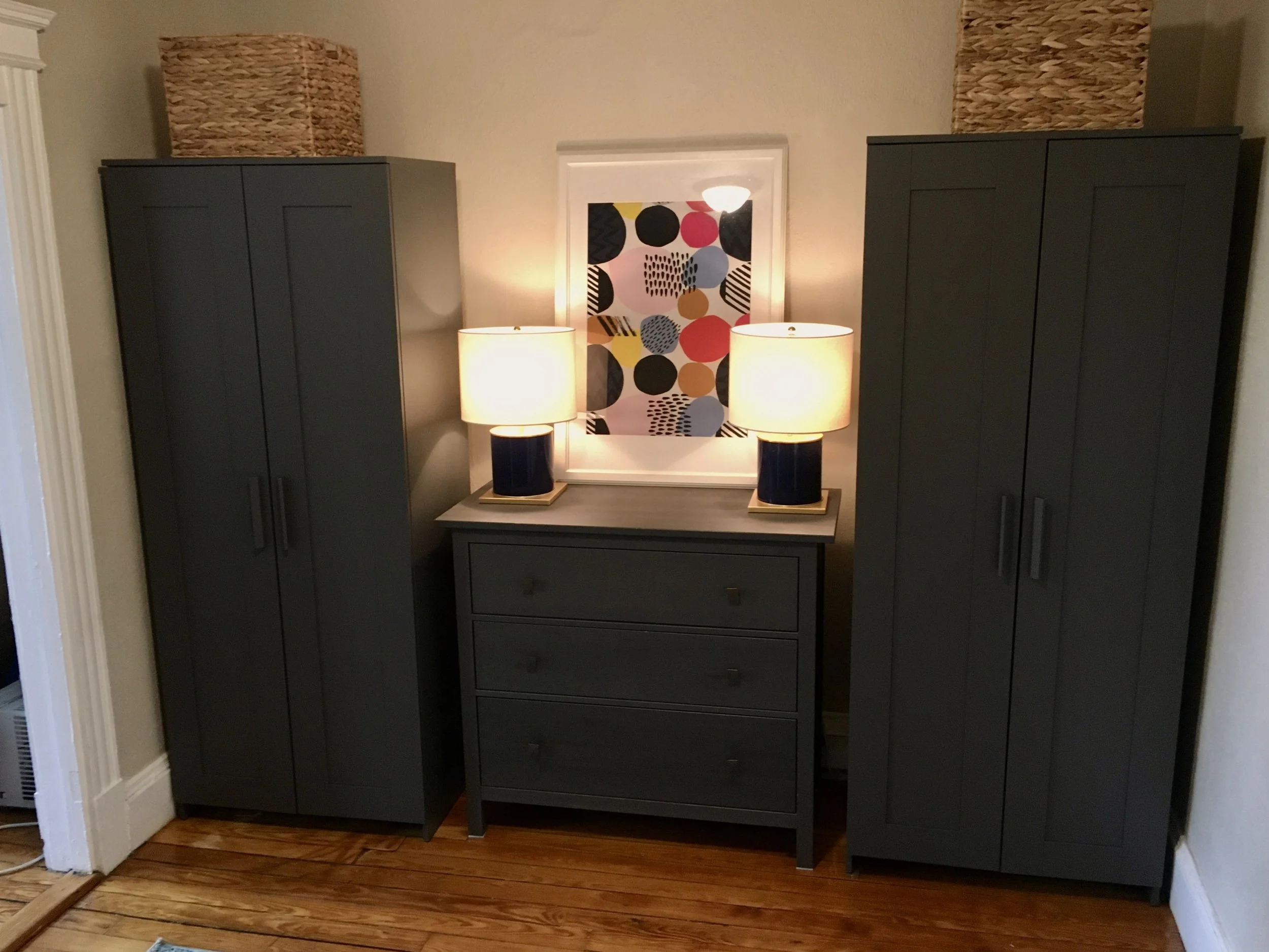 Small apartment storage setup featuring tall wardrobes, woven basket storage, and a coordinating dresser styled with warm lighting for a clean, organized look.