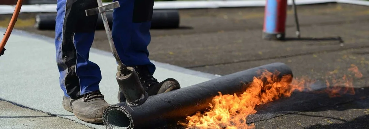 Worker installing a new black roofing membrane on a flat roof while fire is used to heat the material for sealing.