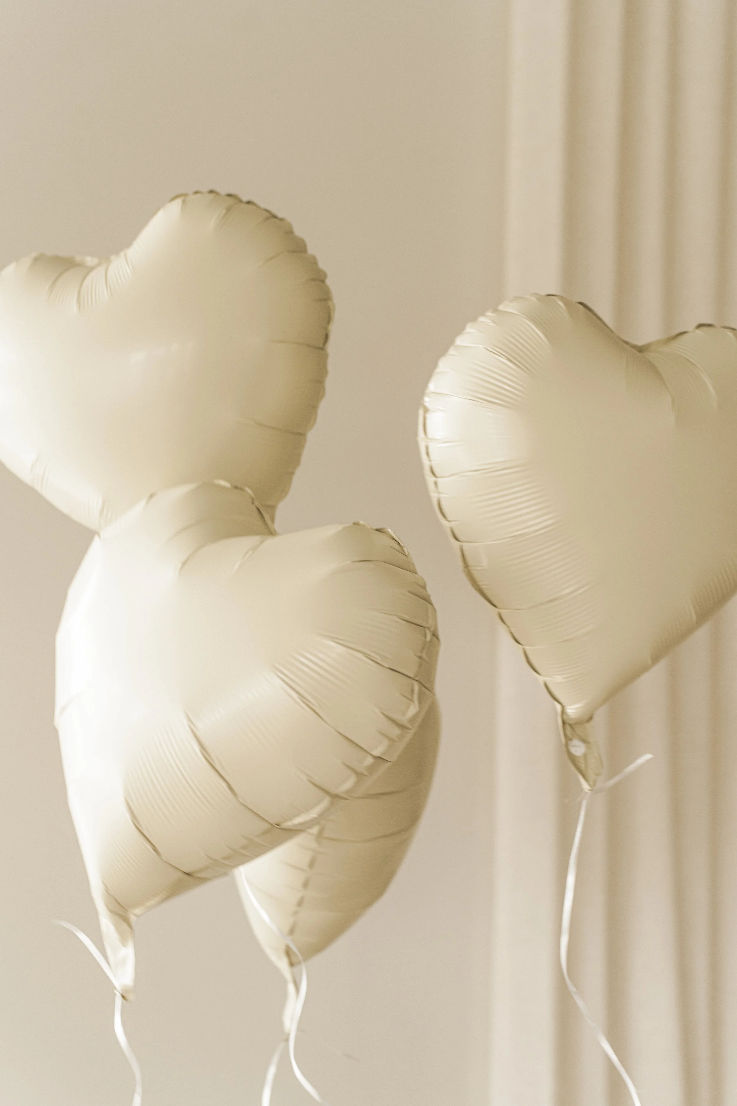 Four cream-colored, heart-shaped balloons float gently indoors against a soft beige background, creating a serene and romantic atmosphere.