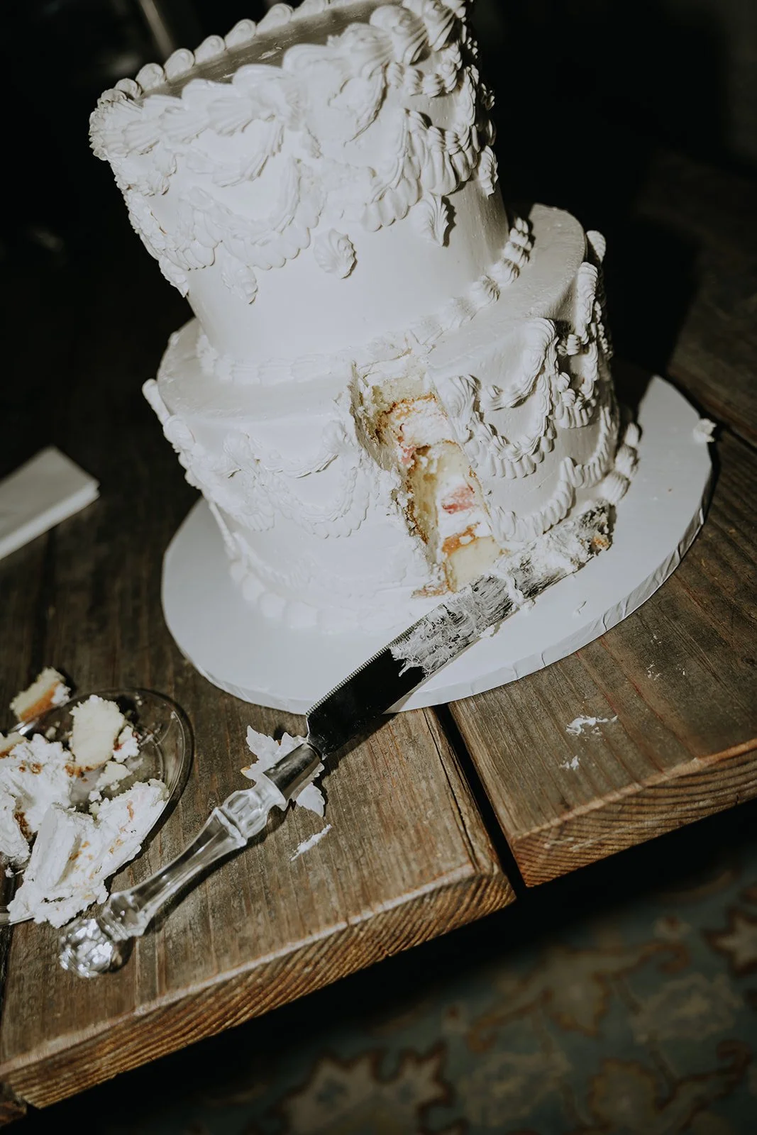 Two-tiered white cake with intricate icing designs, a slice removed revealing layers. Cake knife rests on wooden table with crumbs and a small plate.