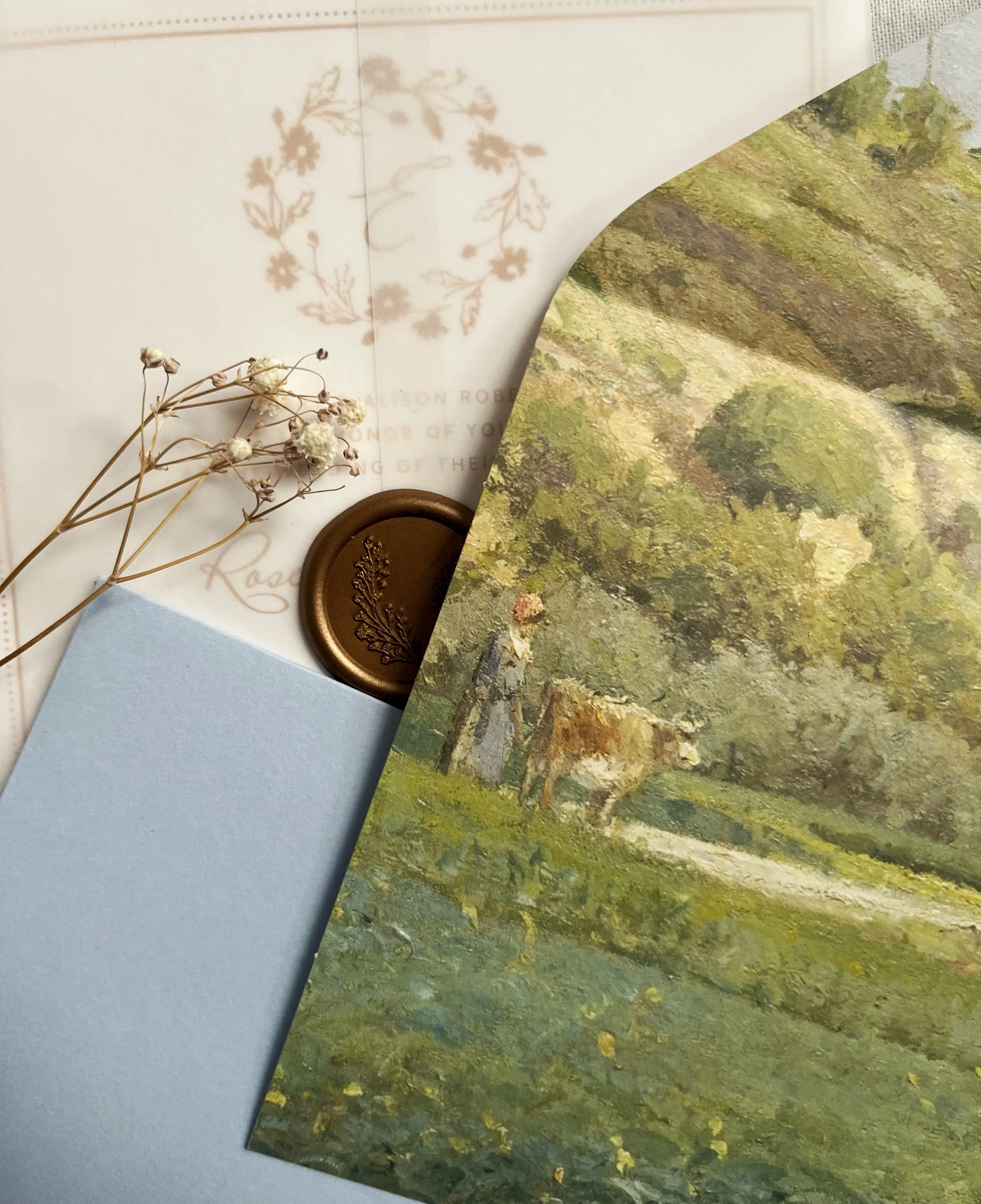 A close-up of a vintage-style envelope with a pastoral painting, featuring a person and a cow. A delicate dried flower and wax seal add elegance.
