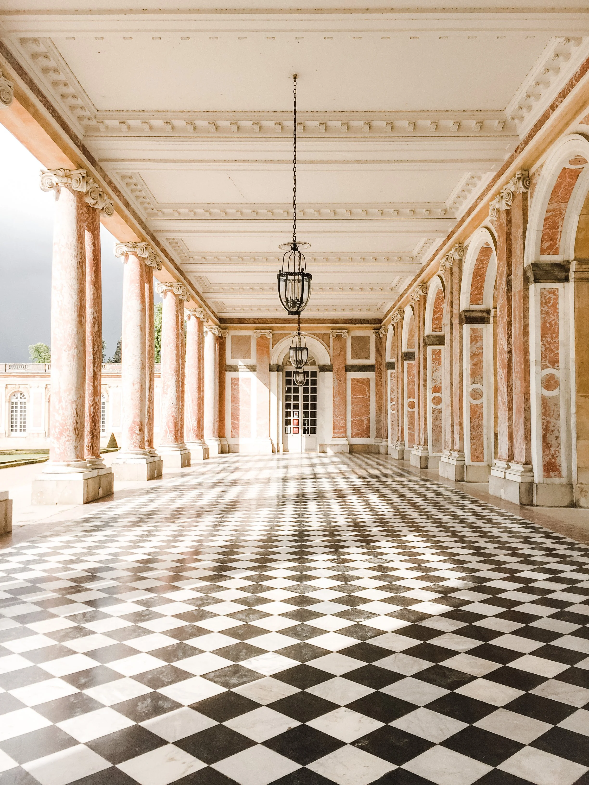 Majestic hallway with pink marble columns, ornate ceiling, and black-and-white checkered floor. Hanging lanterns add elegance under a soft light.