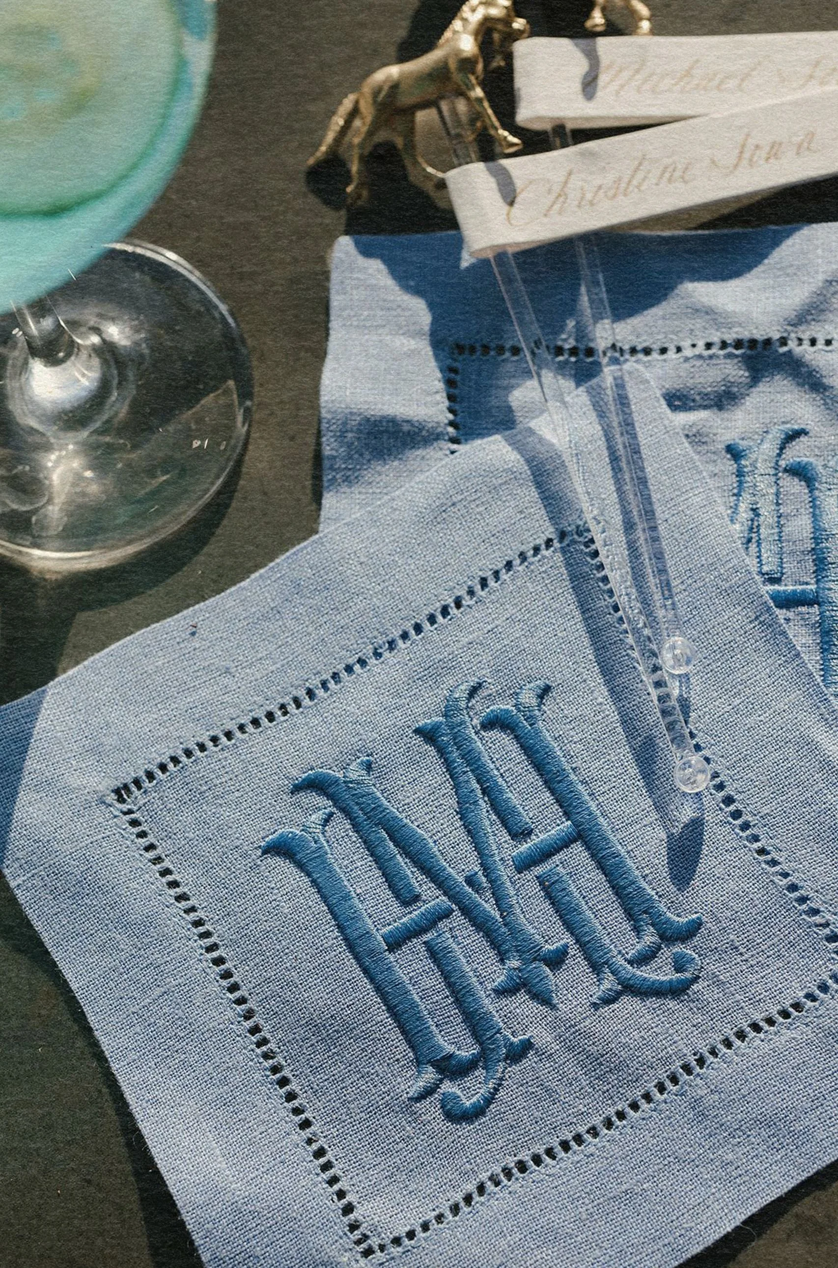 A monogrammed blue napkin with the letters "MH" lies beneath a glass of blue liquid. Nearby, two name cards and a gold animal figurine rest on a dark surface.