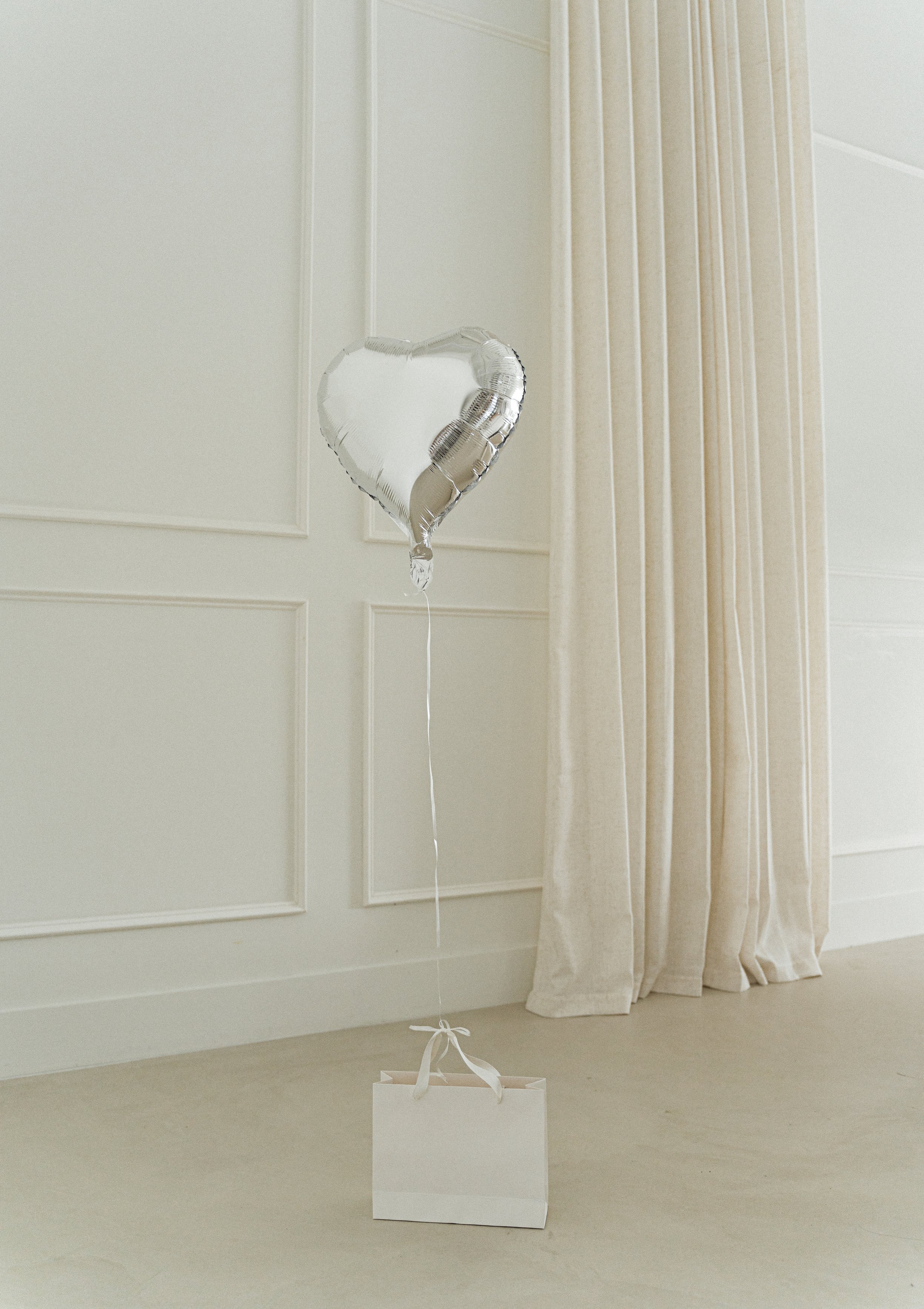 A heart-shaped silver balloon tied to a white gift bag with ribbon on a beige floor. Soft, elegant ambiance with a neutral-colored curtain backdrop.