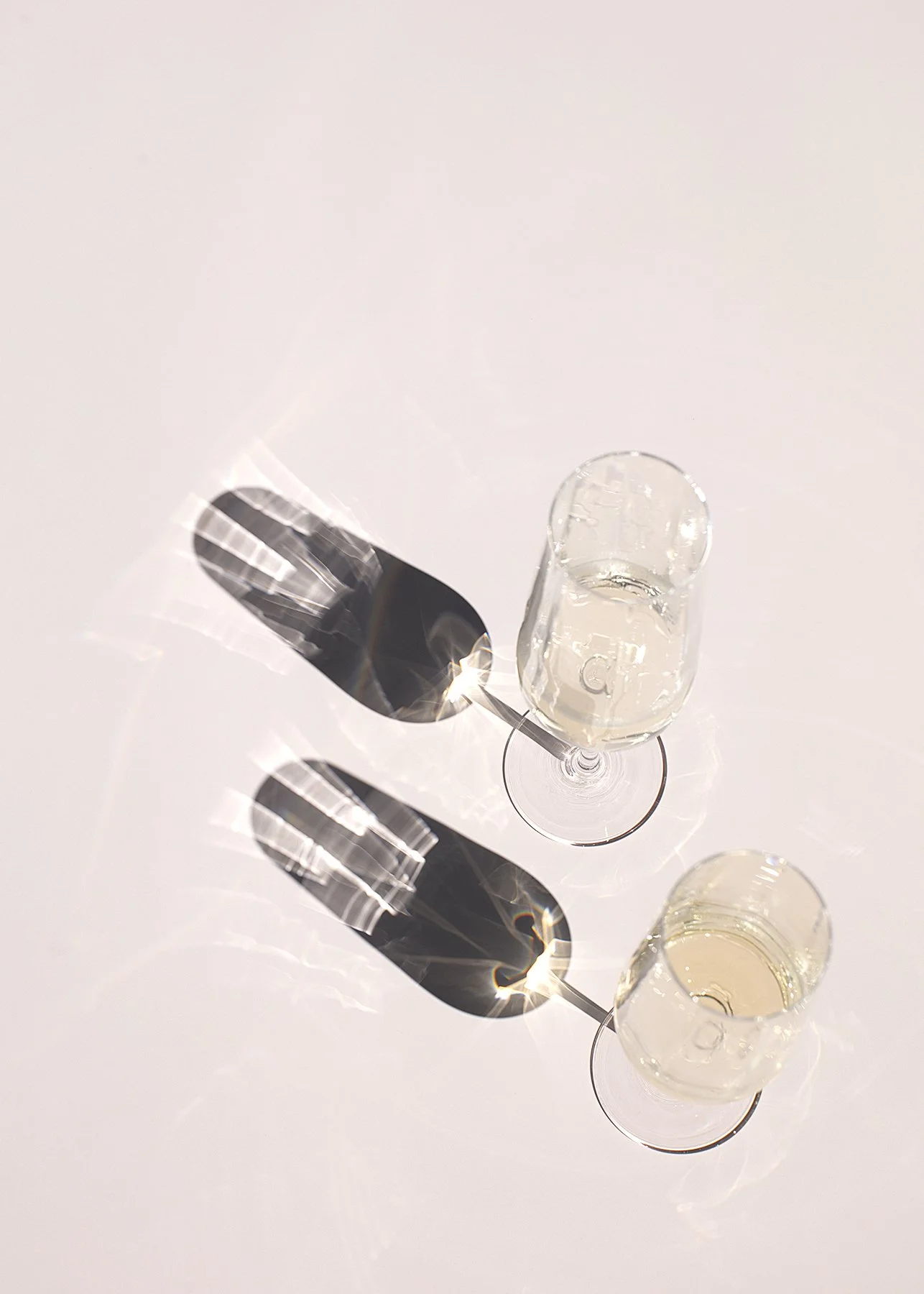 Two champagne flutes casting elongated shadows on a light pink surface. The glasses are filled with pale, sparkling liquid, creating a refined, elegant atmosphere.