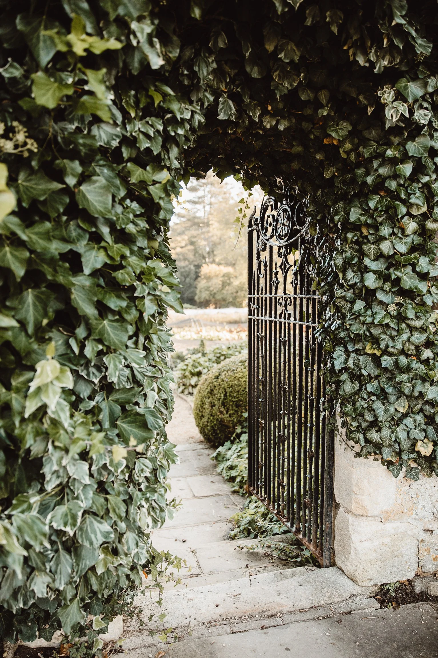A wrought iron garden gate is partially open, framed by lush green ivy. Beyond, a sunlit garden path and manicured bushes evoke a sense of mystery and tranquility.