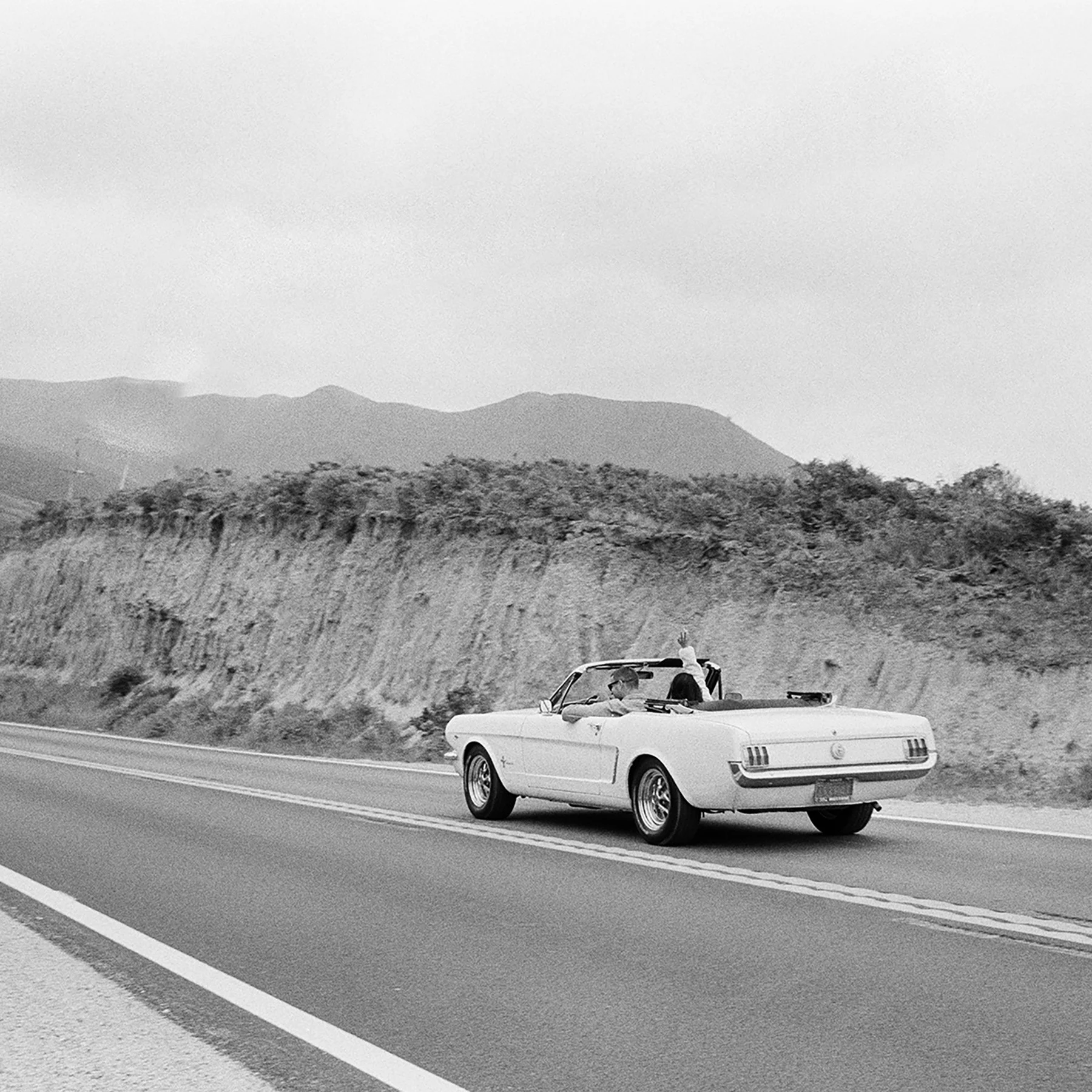 A black-and-white photo of a classic convertible driving down a coastal highway. The car's driver waves joyfully, with cliffs and mountains in the background.