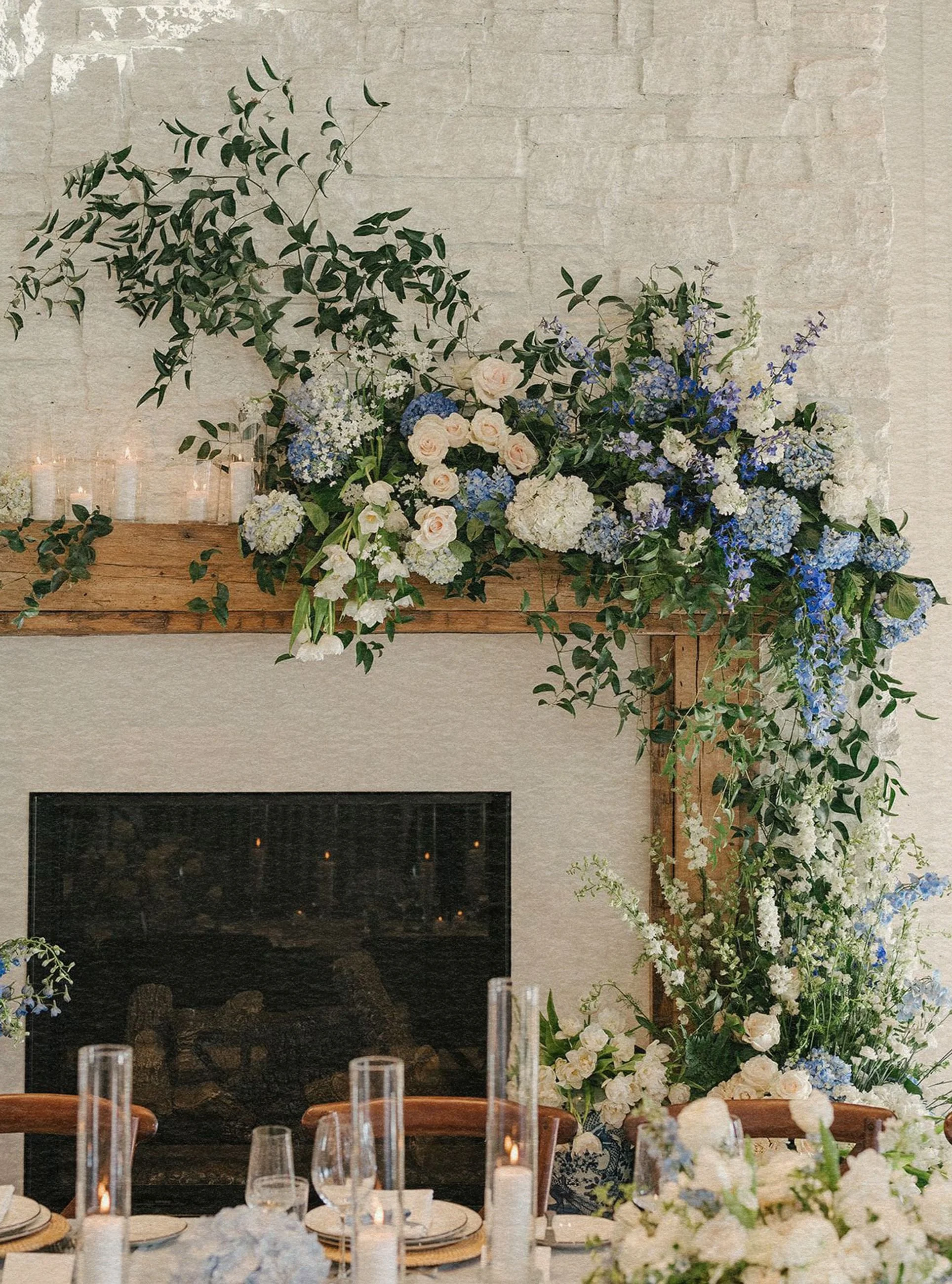 Elegant floral arrangement of blue and white flowers with green foliage on a wooden mantel against a light brick wall, evoking a romantic ambiance.