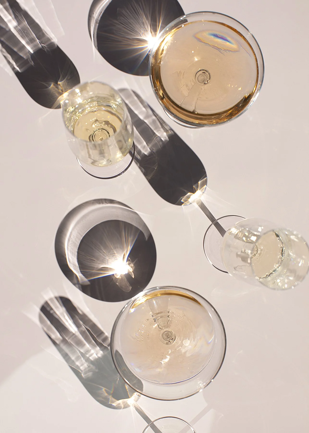 Overhead view of three wine glasses casting intricate shadows and light reflections on a white surface. The scene has a calm and elegant tone.