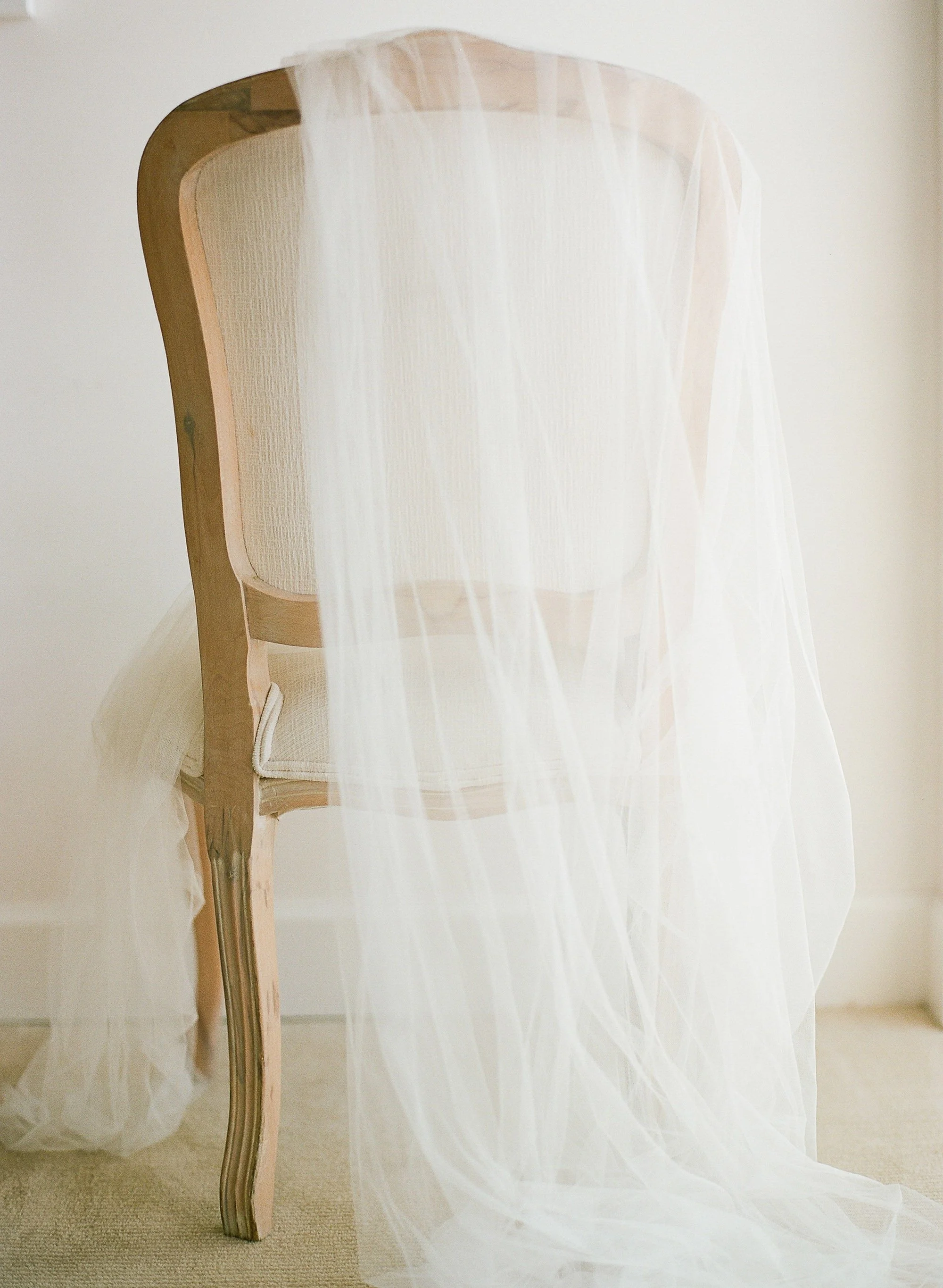 A delicate white veil drapes over the back of a wooden chair against a neutral wall, creating a soft, elegant, and serene atmosphere.