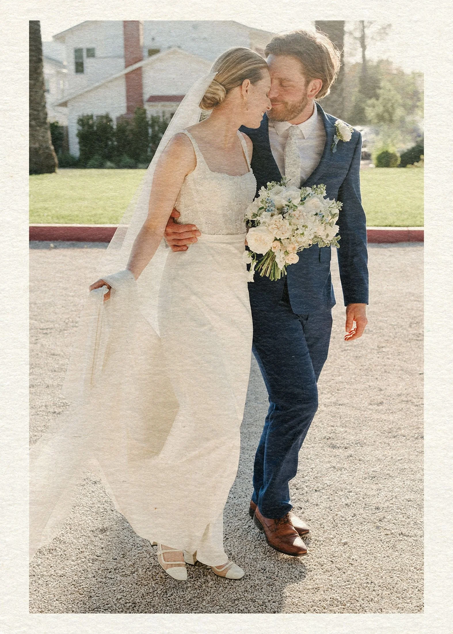 A bride and groom walk arm in arm, smiling warmly. The bride wears a white dress and veil, holding a bouquet, while the groom is in a blue suit.