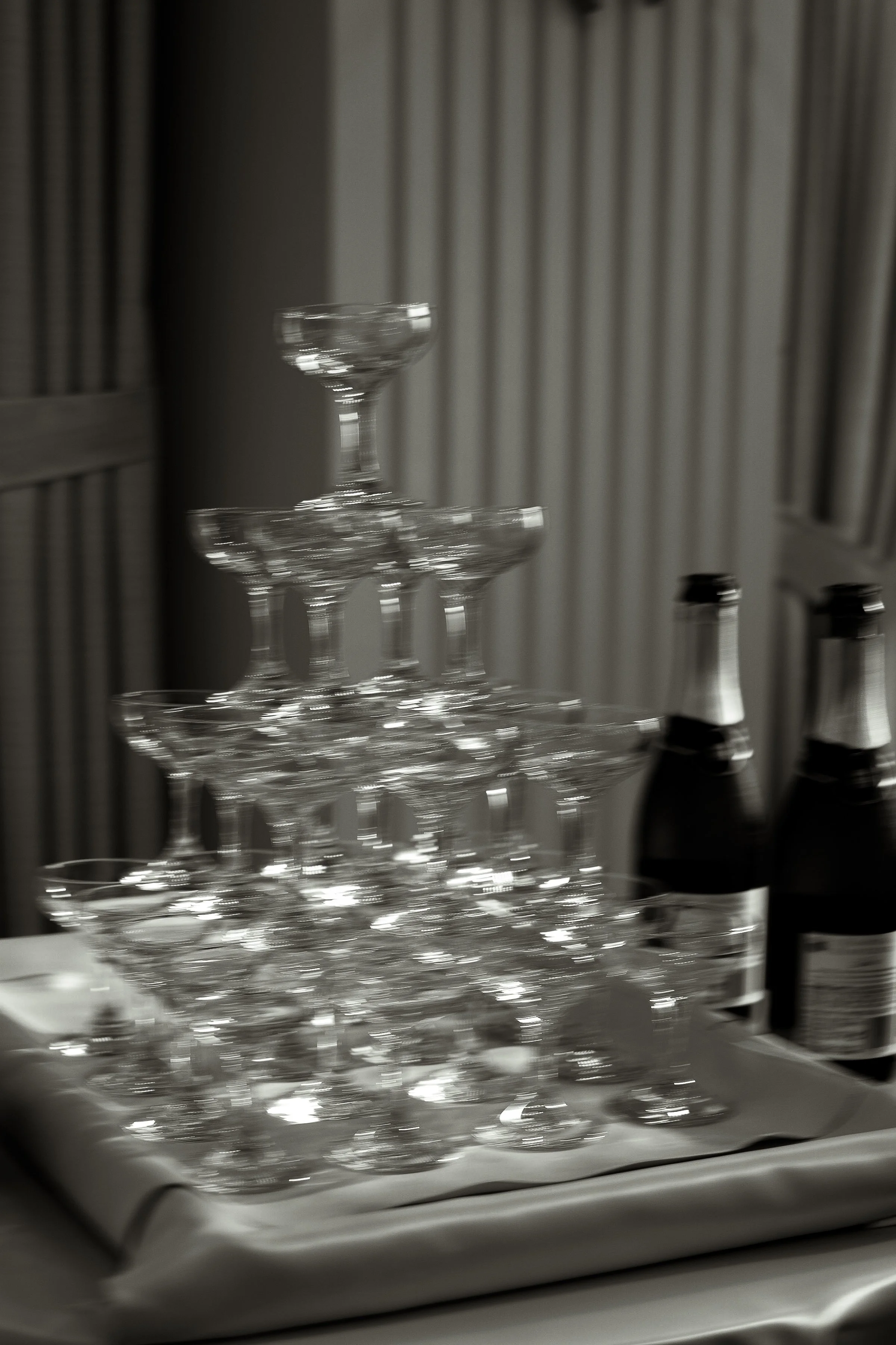 A black and white image shows a pyramid of stacked champagne glasses on a table with two bottles beside them. The setup conveys an elegant, celebratory vibe.