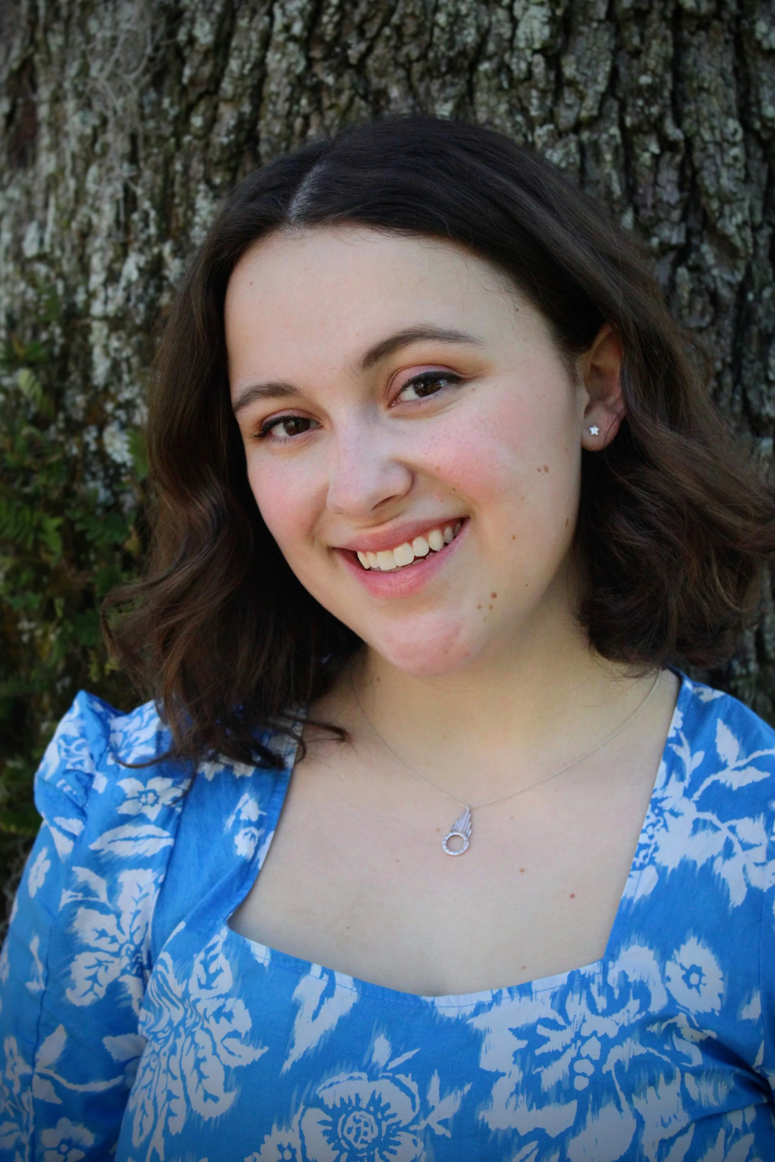 A young woman with wavy brown hair, wearing a blue floral dress and a silver necklace, smiling outdoors in front of a tree bark background.