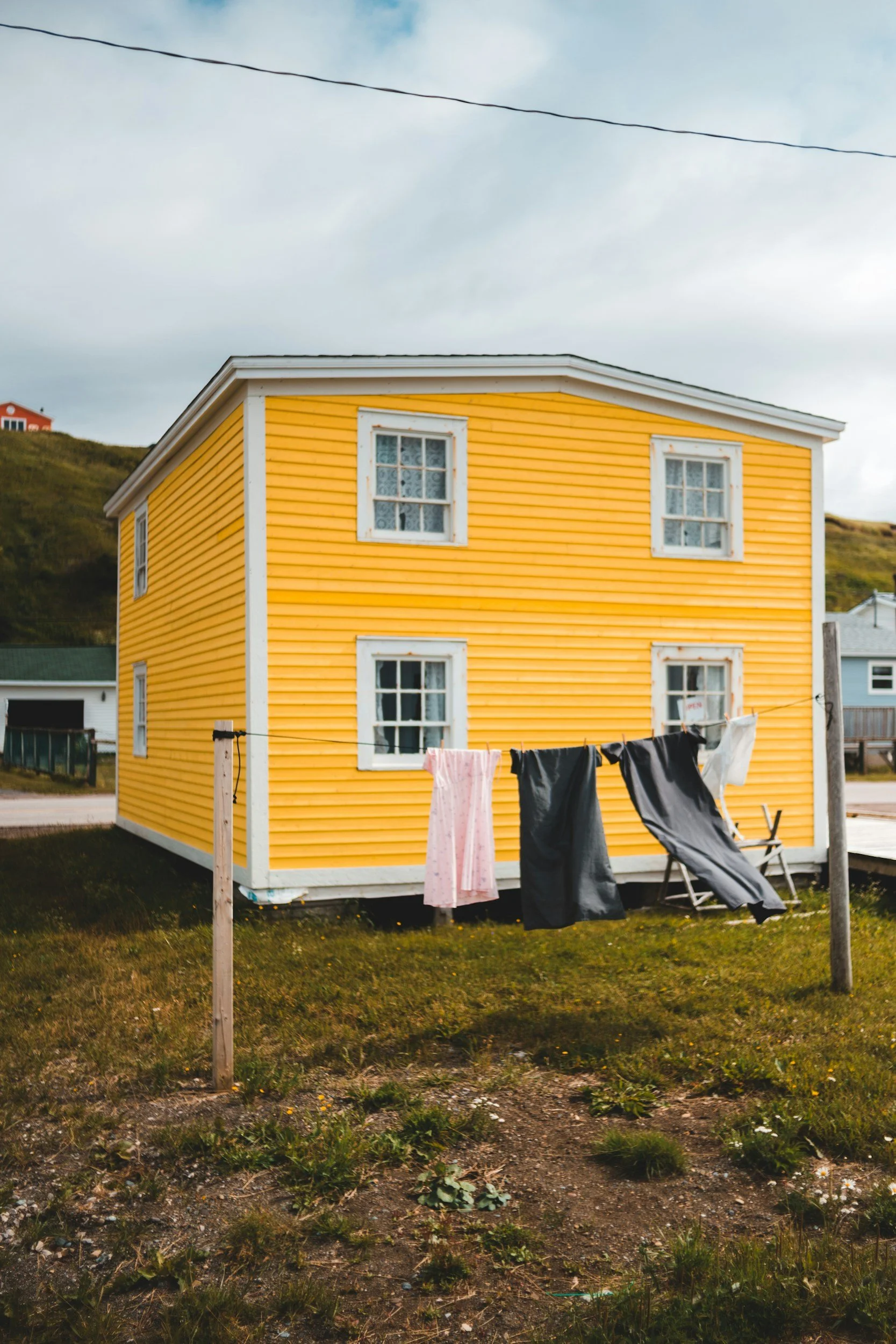 A two-story yellow house with white window frames and a clothesline with clothing hanging outside. The sky is cloudy and there are hills in the background.