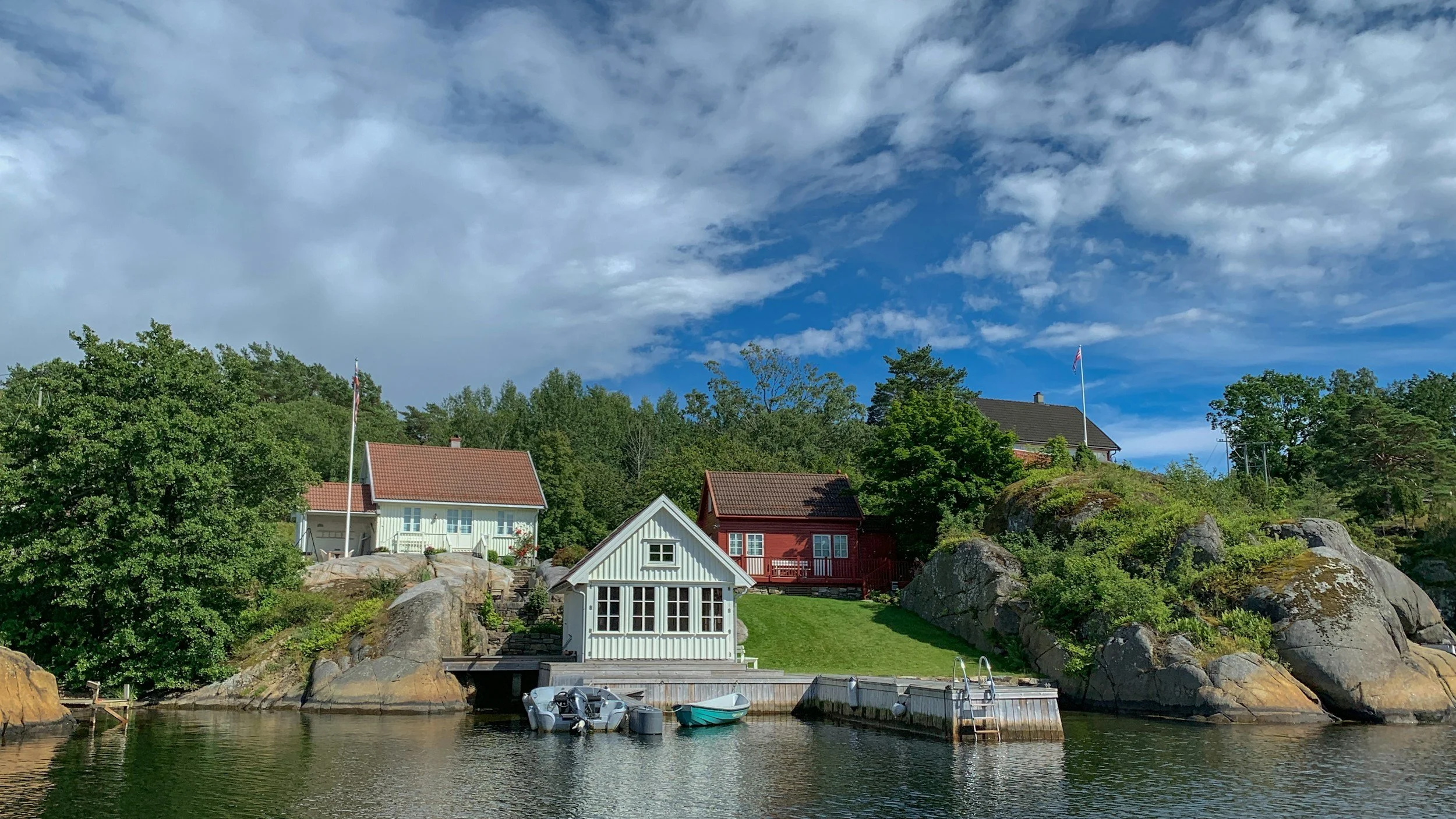 A Scandinavian-style lakeside scene with houses on a hill, boats docked at a small pier, greenery, rocks, and a partly cloudy blue sky.