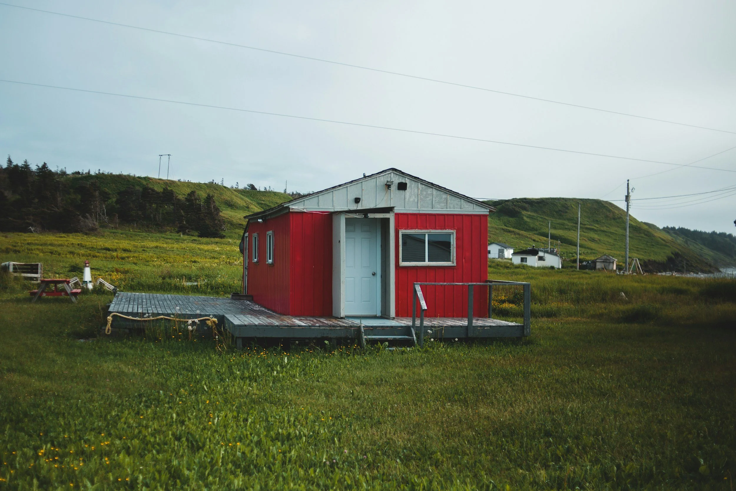 Red small house on a wooden deck situated in a grassy field, with green hills and a cloudy sky in the background.