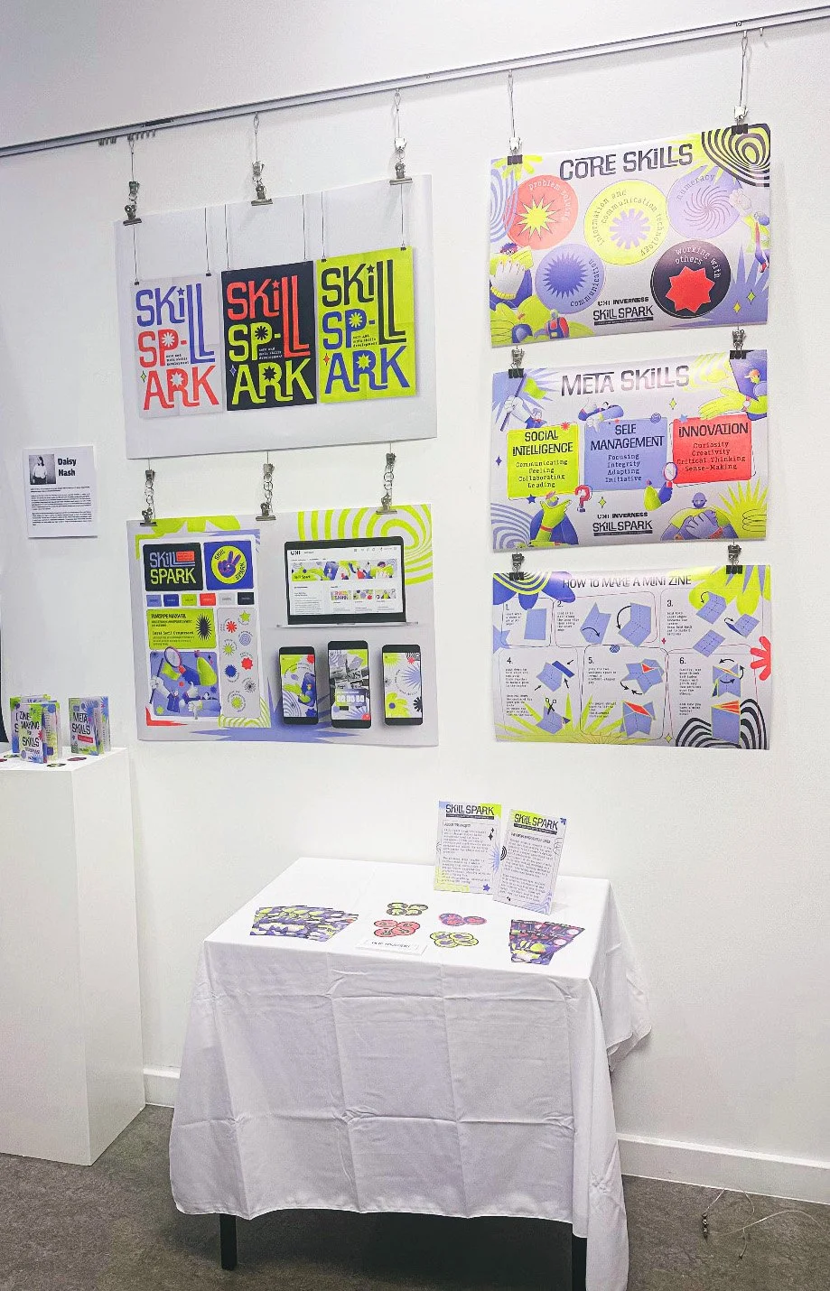 Display of educational posters and materials about SkillSpark, a learning platform, on a white wall with a table featuring brochures and small paper models.