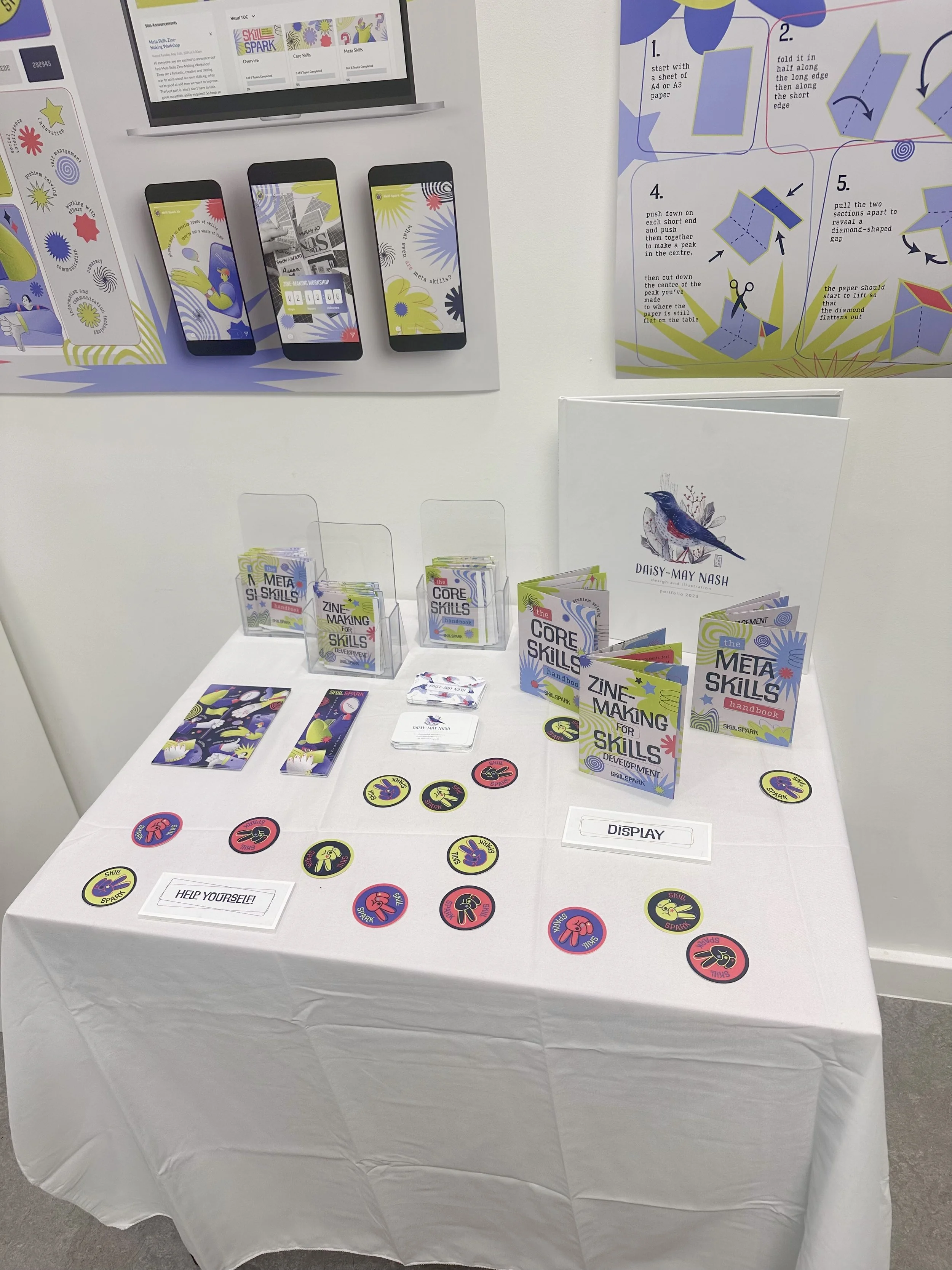Display table with educational materials and promotional items for 'Core Skills' and 'Zine Making for Skills Development,' including booklets, circular badges, and pamphlets, with informational signage and posters behind.