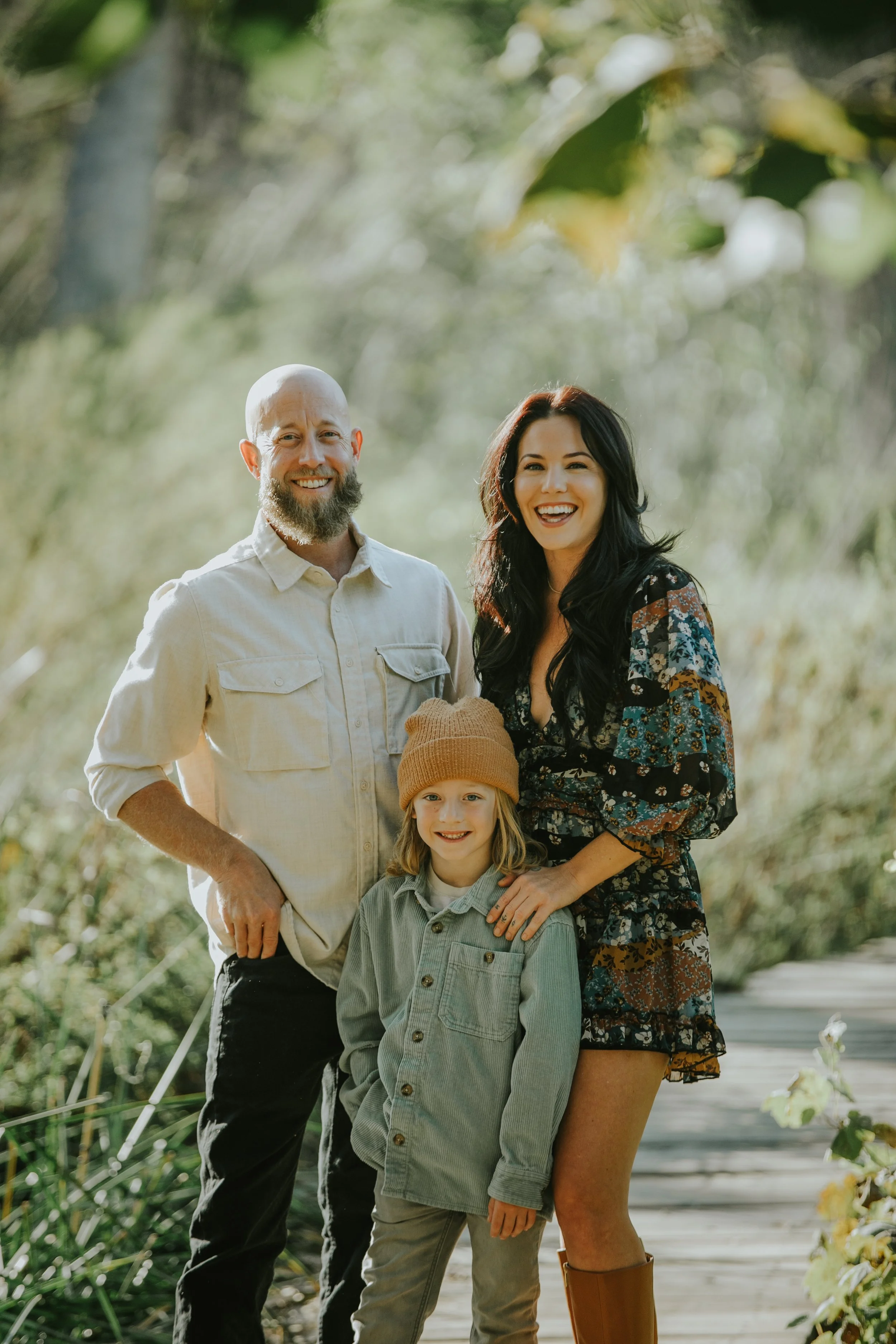 A family of three standing outdoors on a wooden path in a lush, green environment. The man has a beard and is wearing a light-colored shirt. The woman has long dark hair and is wearing a dark floral dress. The child, a girl, has blonde hair, a beige knit hat, and a green jacket.