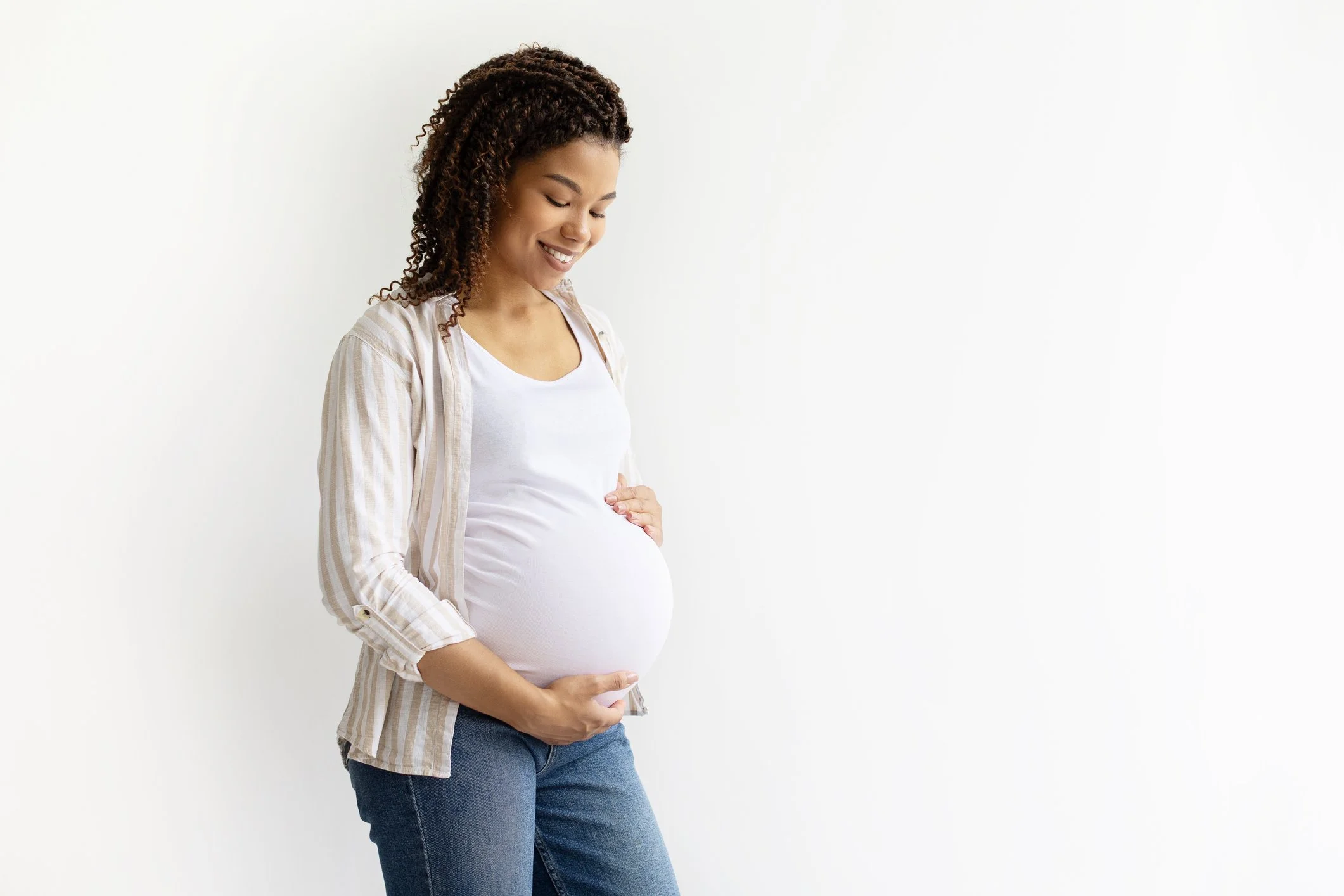 Smiling pregnant woman holding her baby bump while standing, expressing happiness and well-being.