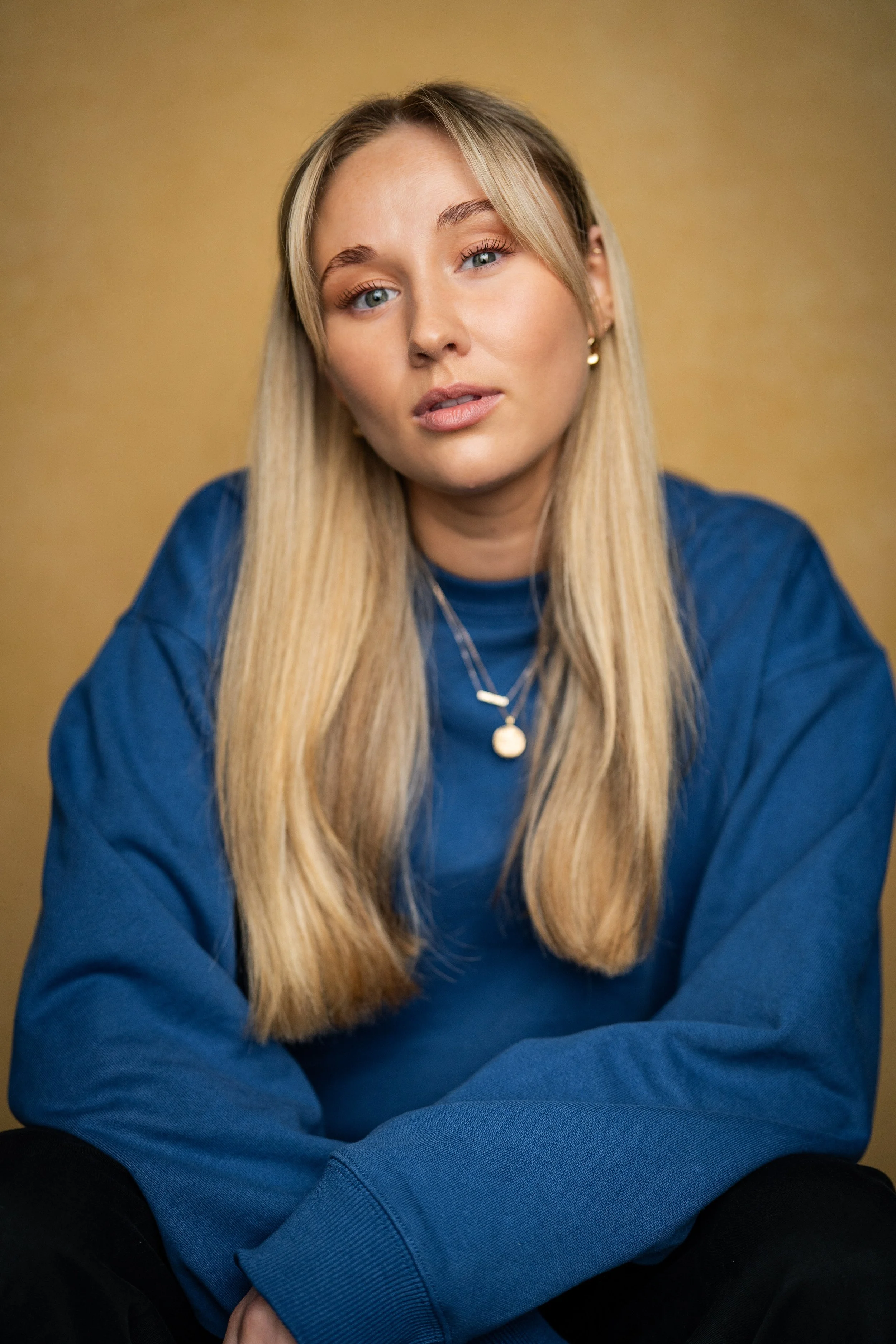 A young woman with long blonde hair, blue eyes, wearing a blue sweatshirt, gold hoop earrings, and layered necklaces, sits against a yellow background.