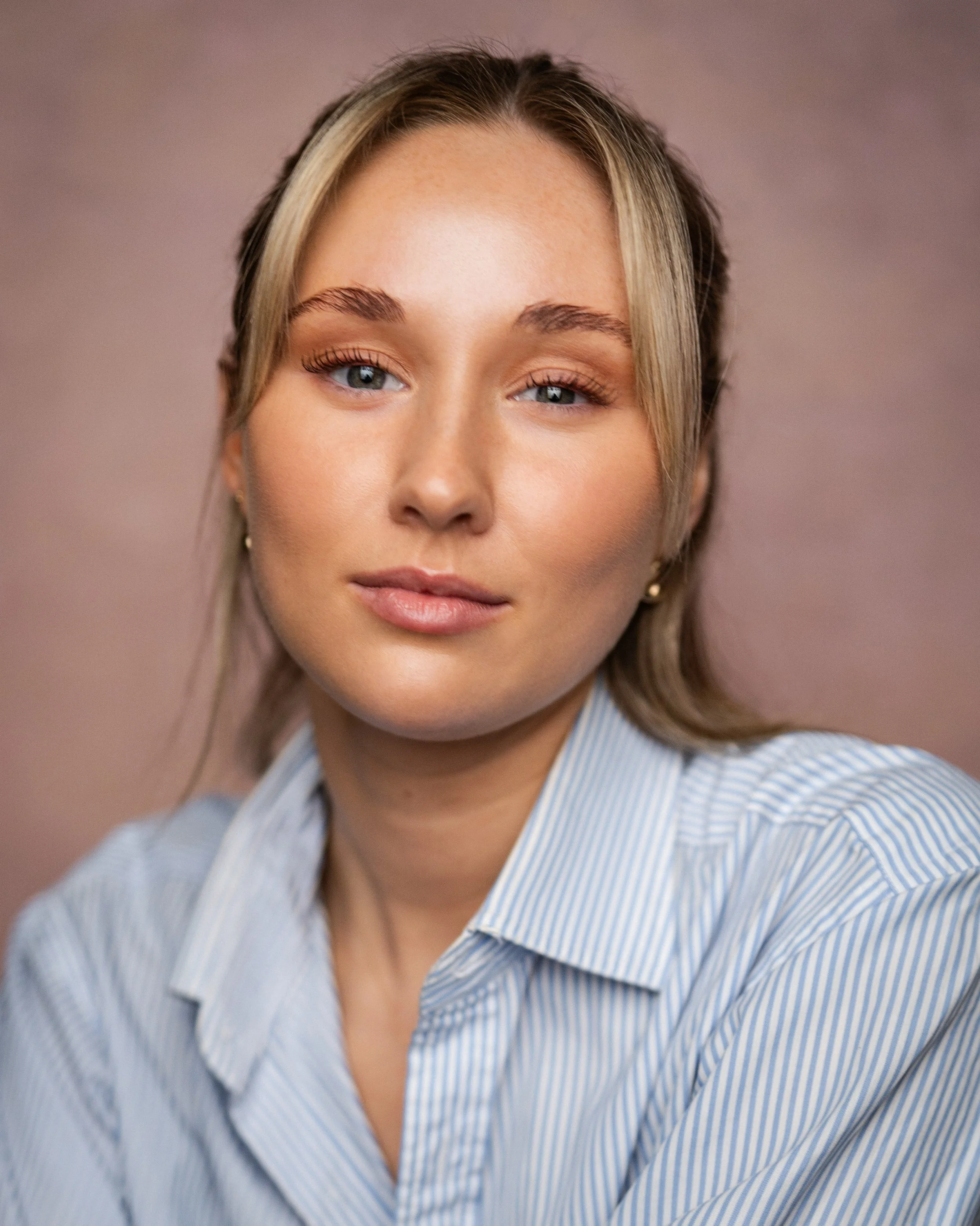 A close-up portrait of a woman with blonde hair, blue eyes, and light skin, wearing a light blue striped shirt against a neutral background.