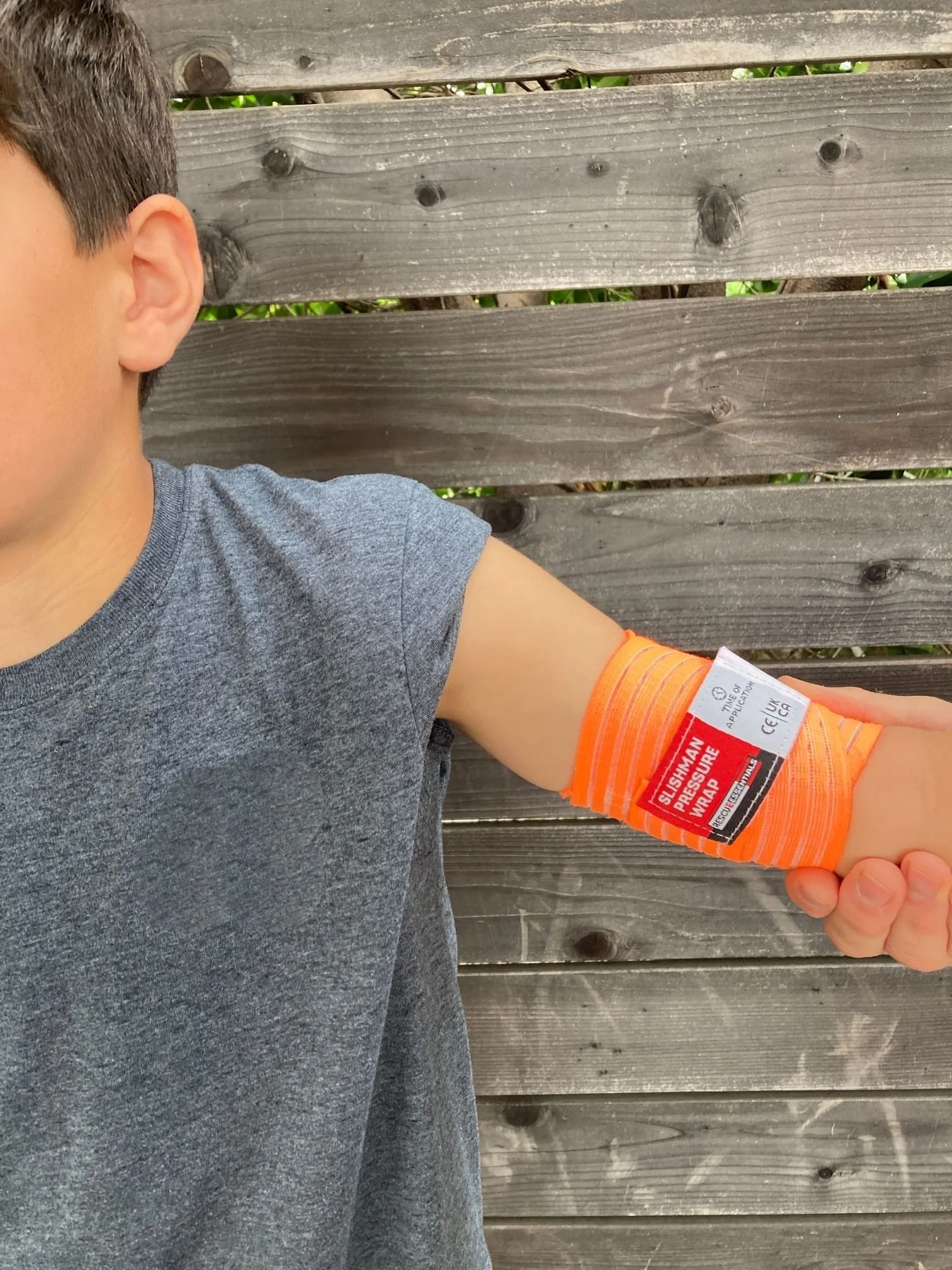 A young boy is showcasing a blood pressure cuff wrapped around his upper arm. The cuff is orange with a label indicating it is a silkiman pressure wrap. The boy is wearing a gray T-shirt and standing in front of a wooden fence.