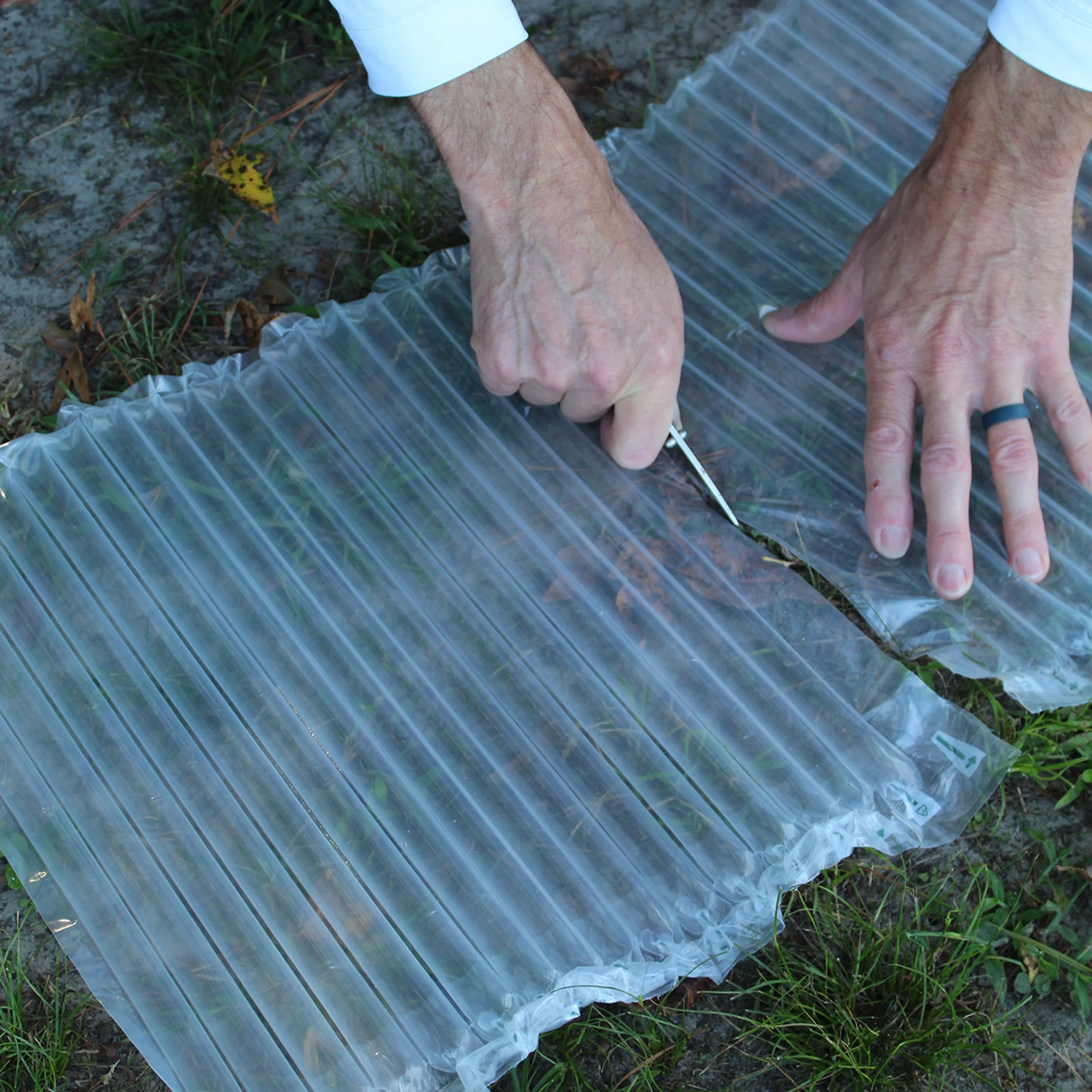 Person cutting open a clear plastic air cushion with a small knife on the ground outdoors.