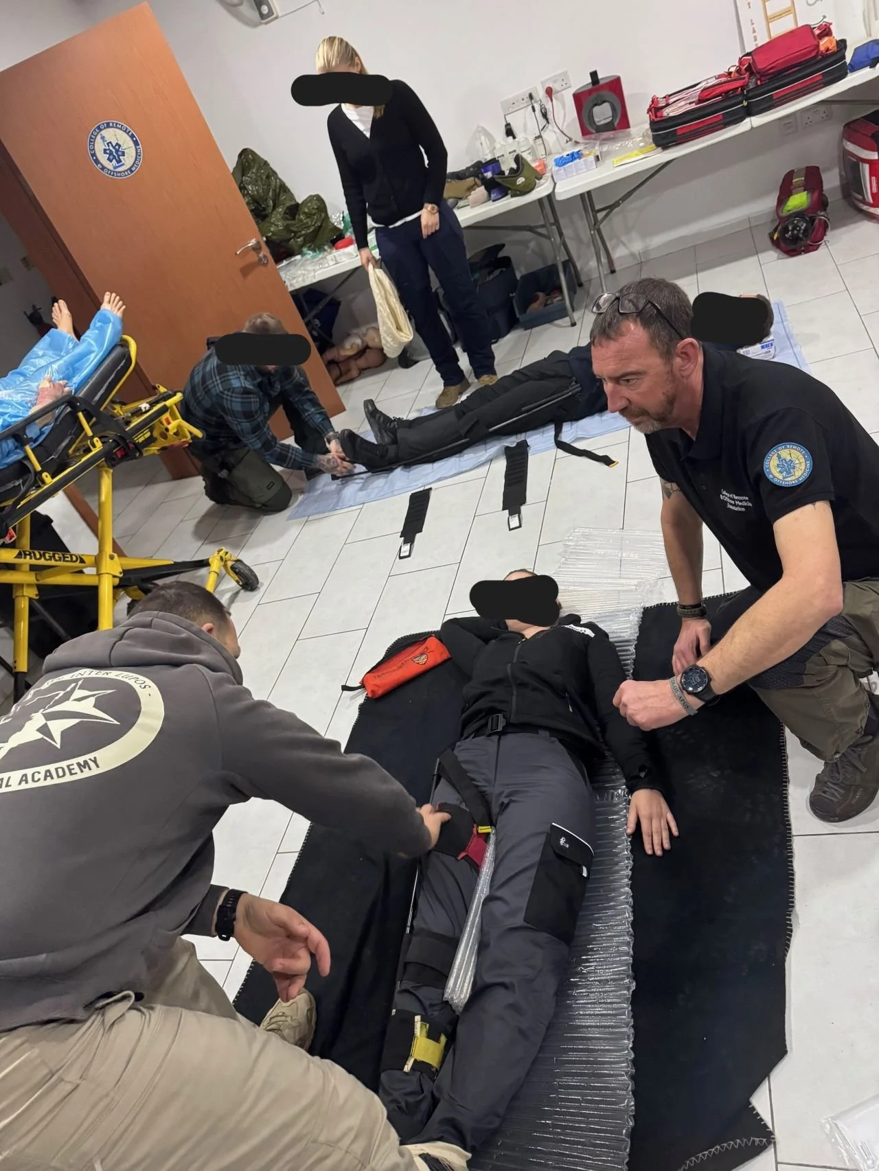 Medical personnel providing emergency care to a person lying on a stretcher. Several people assisting, with medical supplies and equipment in the background, in a clinical setting.