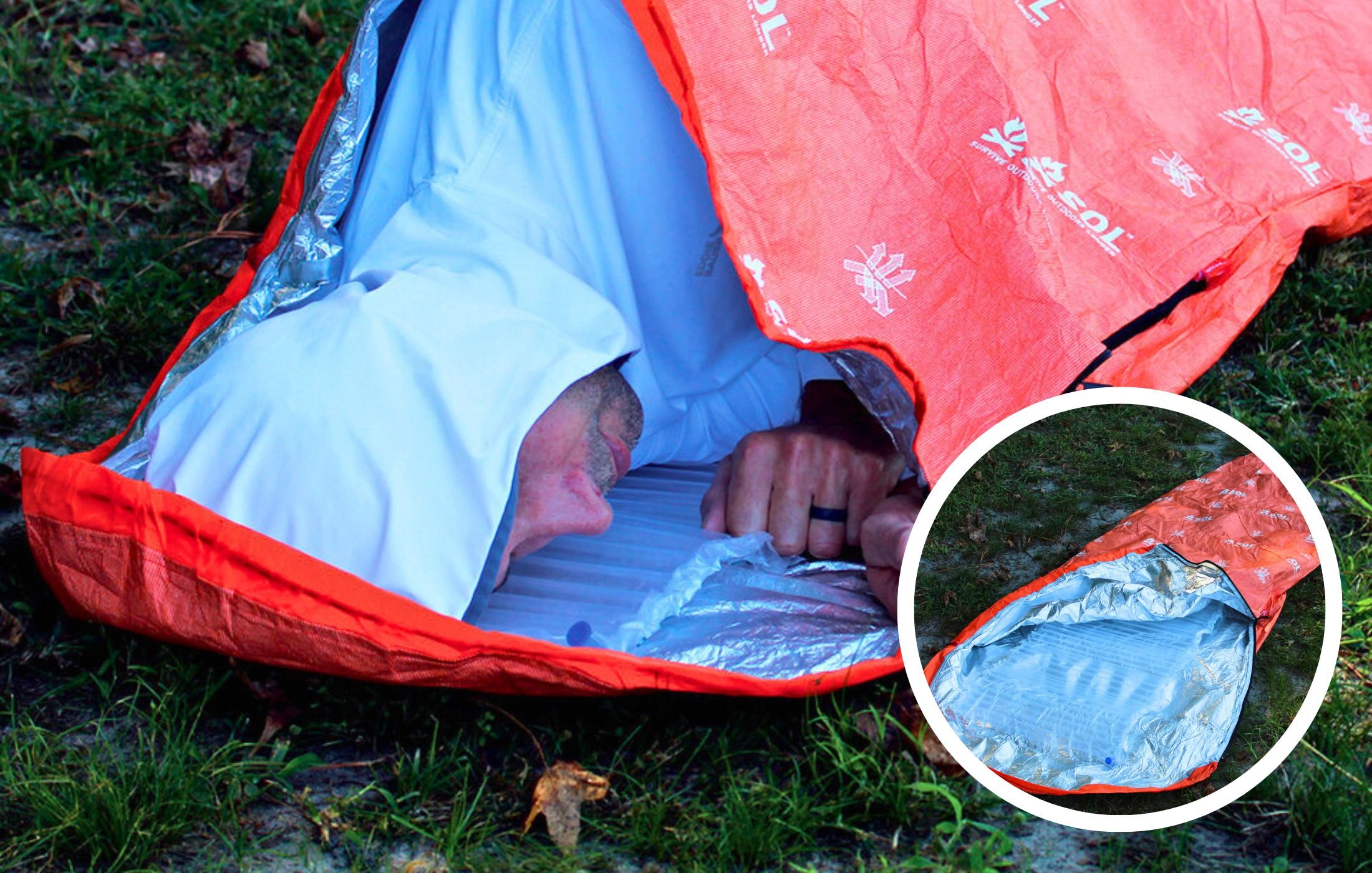 A person in a white shirt and black ring lying inside an orange and silver emergency blanket, partially covered by an orange emergency tent on grass, seen from an angle with a focus on their head and clenched fist.