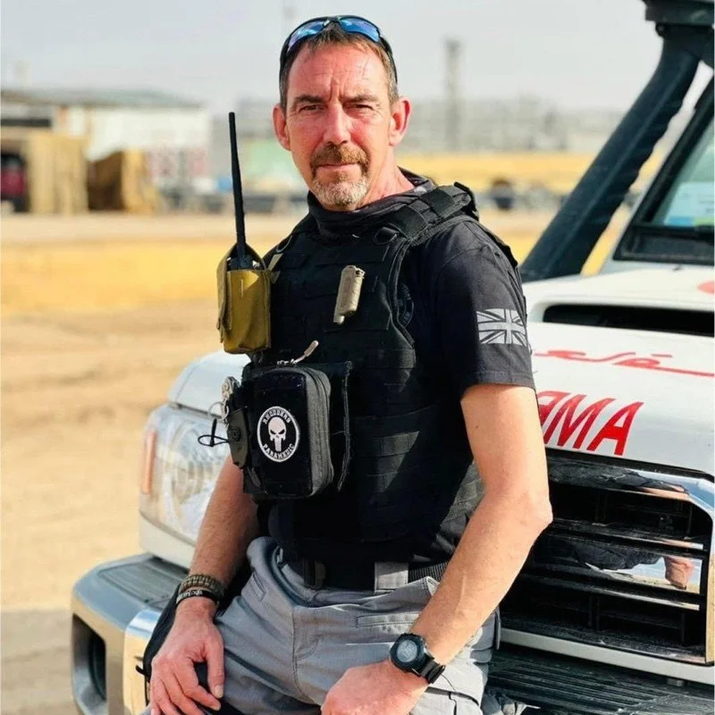 A middle-aged man with a beard and sunglasses resting on his head, standing outdoors in front of an off-road vehicle with ambulance markings. He's wearing tactical gear, including a black vest with pouches, and has a serious expression.