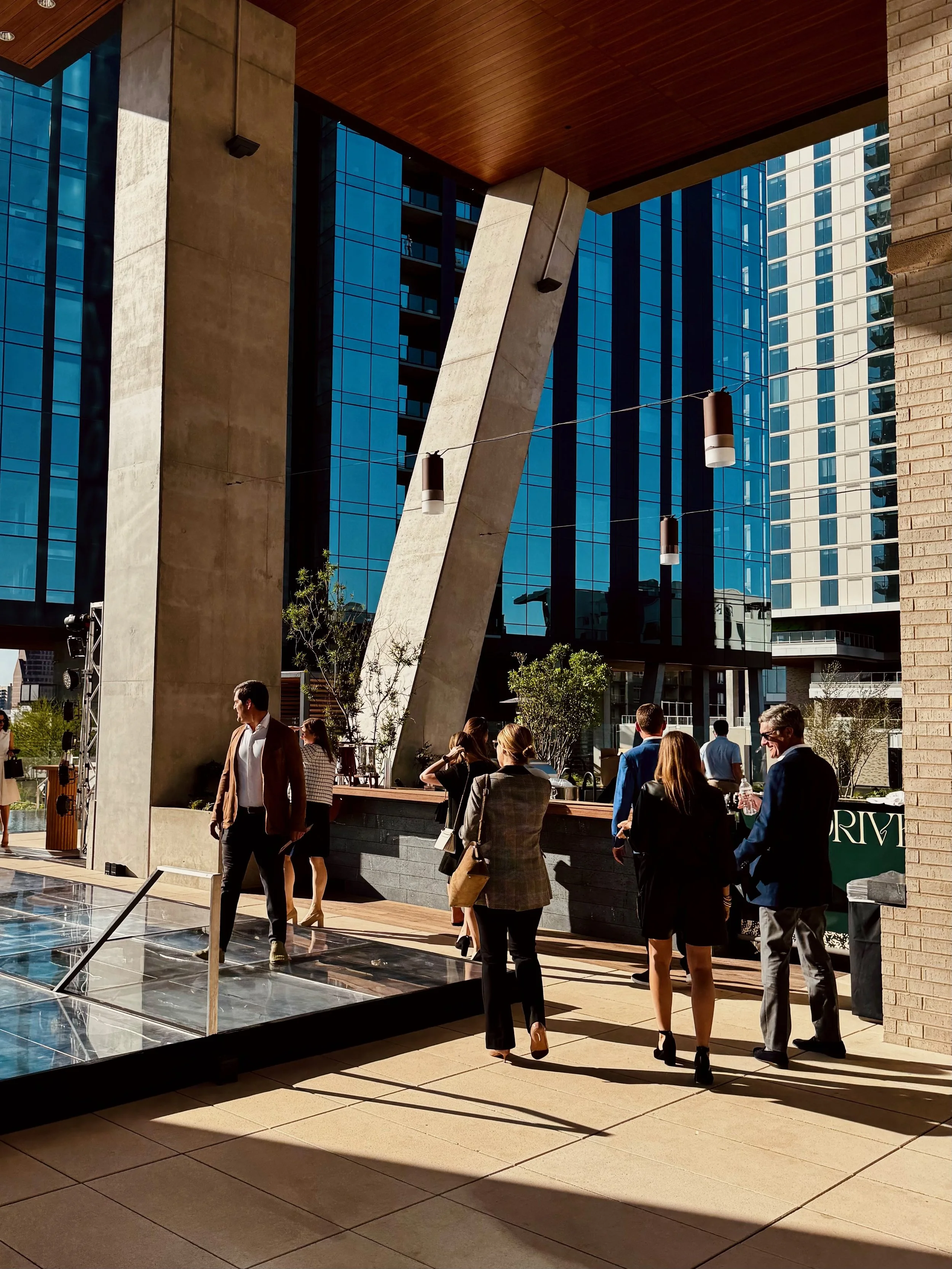 People in business attire walking in an outdoor urban setting with modern buildings, trees, and a reflective glass surface.