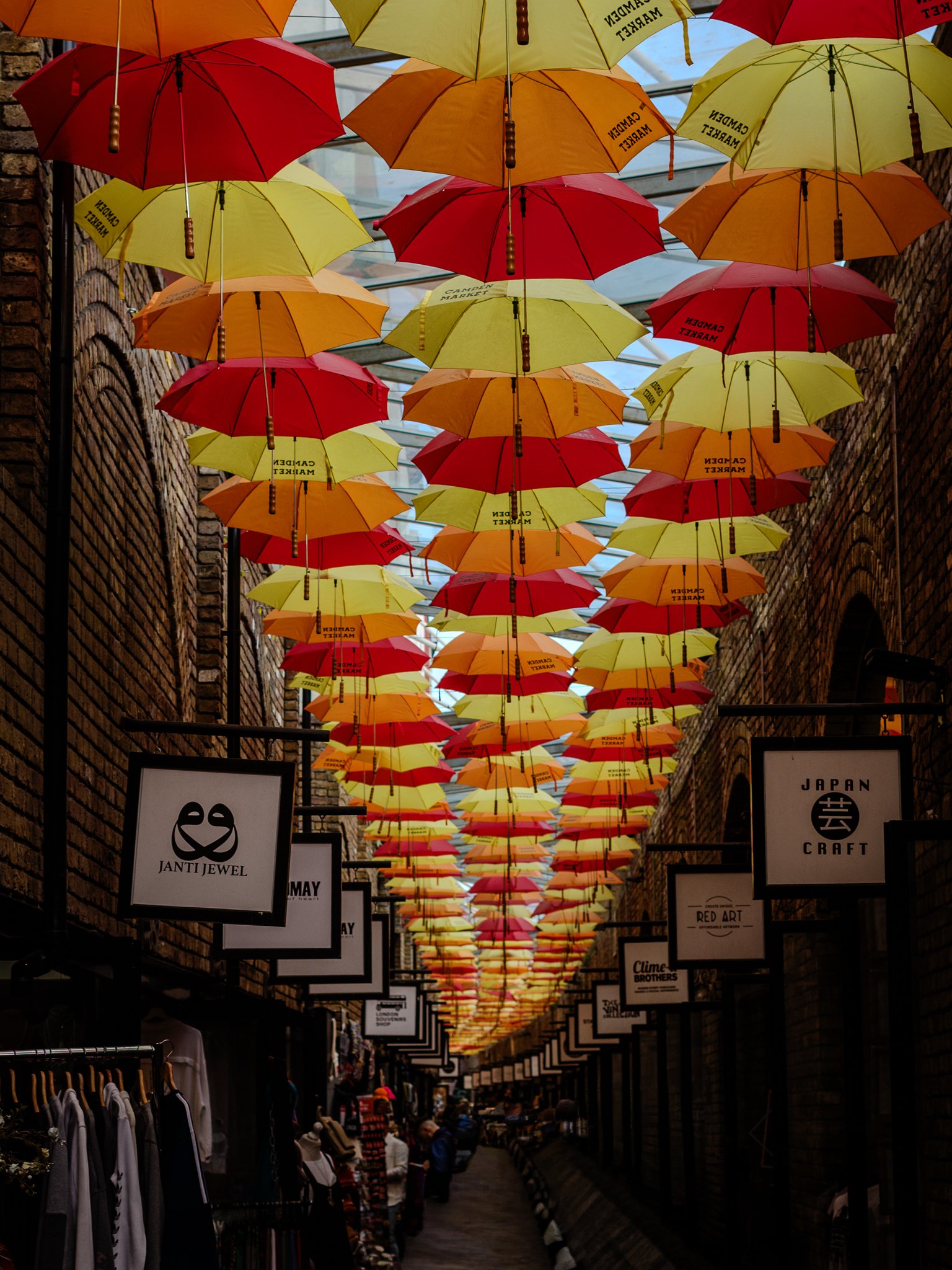 A street decorated with hanging umbrellas in yellow, red, and orange colors, creating a colorful canopy overhead. The street is lined with shops, with signs displaying various store names, and a few people browsing.