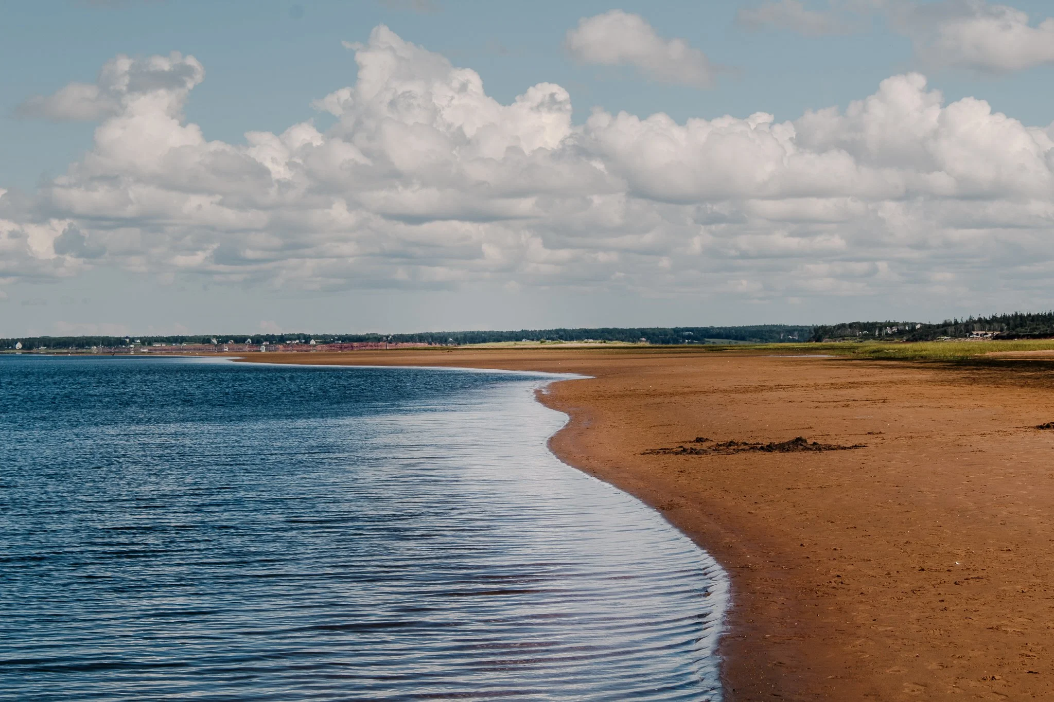 A wide view of a shoreline with a sandy beach, blue water, a cloudy sky, and distant land and buildings in the background.