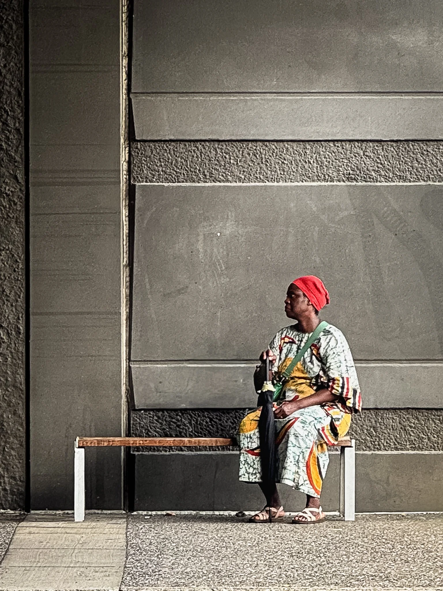 A woman sitting on a wooden bench against a gray wall, wearing a colorful patterned dress, a red hat, and sandals, holding a black bag and a green shoulder strap.